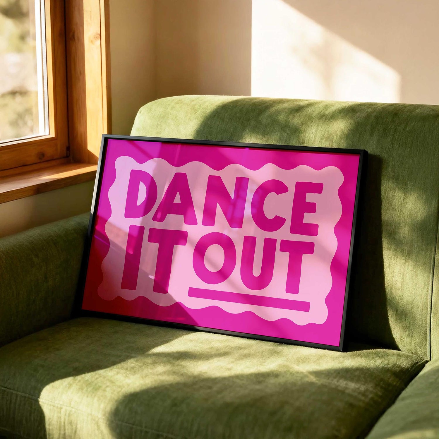 A framed wall art print in hot pink with the text DANCE IT OUT inside a wavy border. The frame is leaning on an olive green sofa by a sunny window.