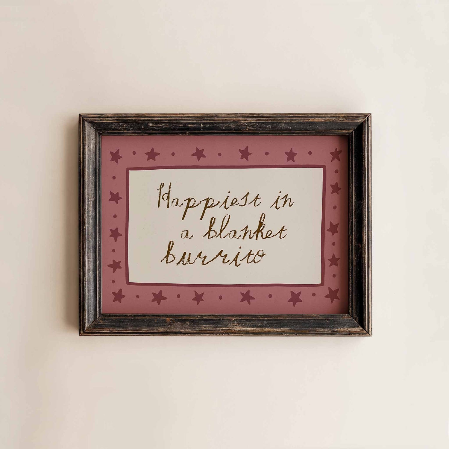 A rustic wooden frame hanging on a white wall. Inside the frame is a whimsical print with a hand drawn star border in dusky pink and the words 'happiest in a blanket burrito' in a child like handwritten font.