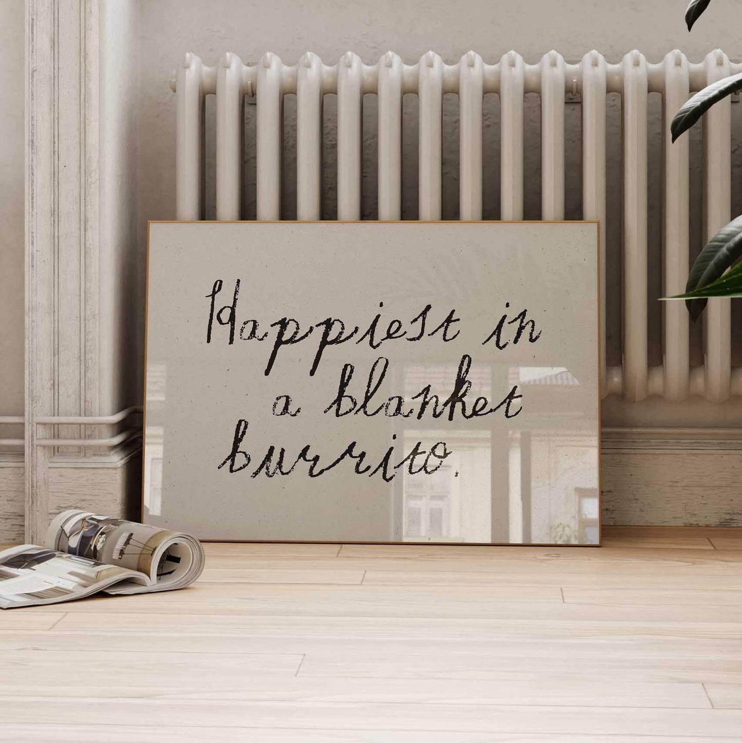 A framed quote on a wall that says "happiest in a blanket burrito" is displayed on a wooden floor next to a radiator.