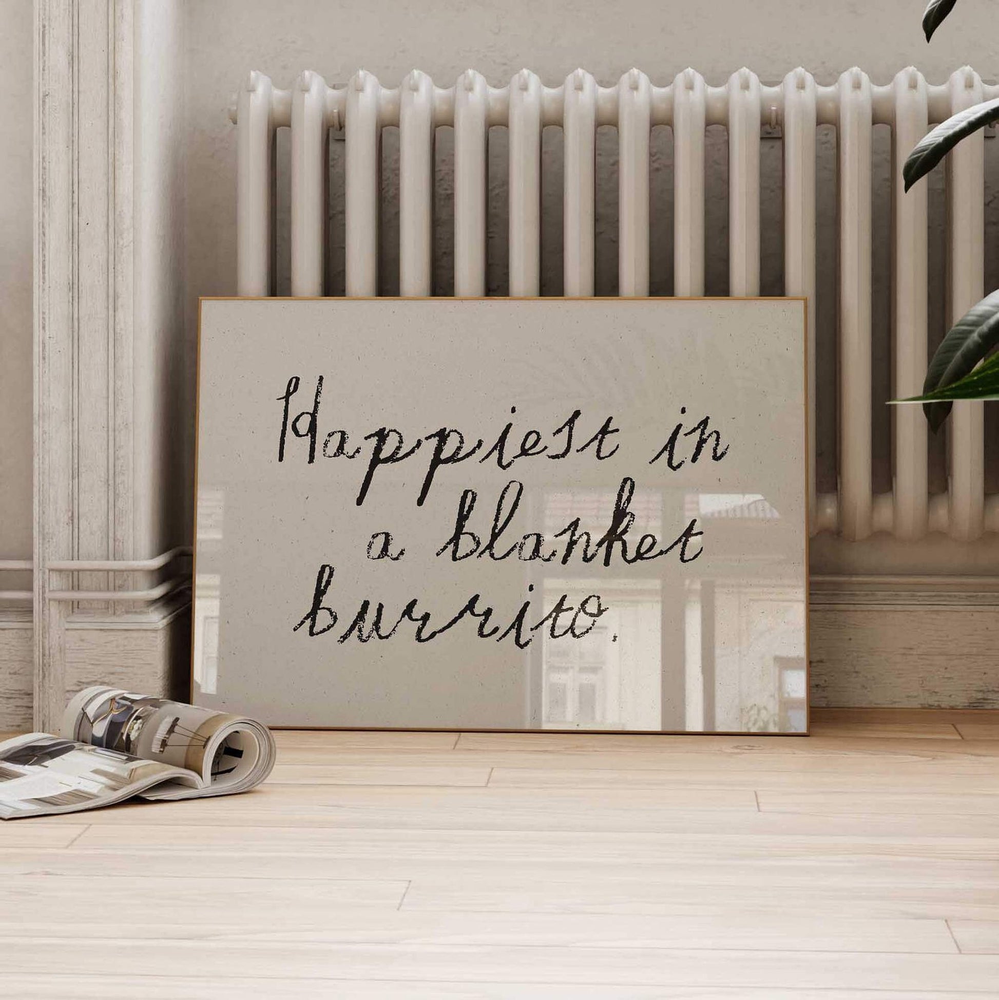 A framed quote on a wall that says "happiest in a blanket burrito" is displayed on a wooden floor next to a radiator.