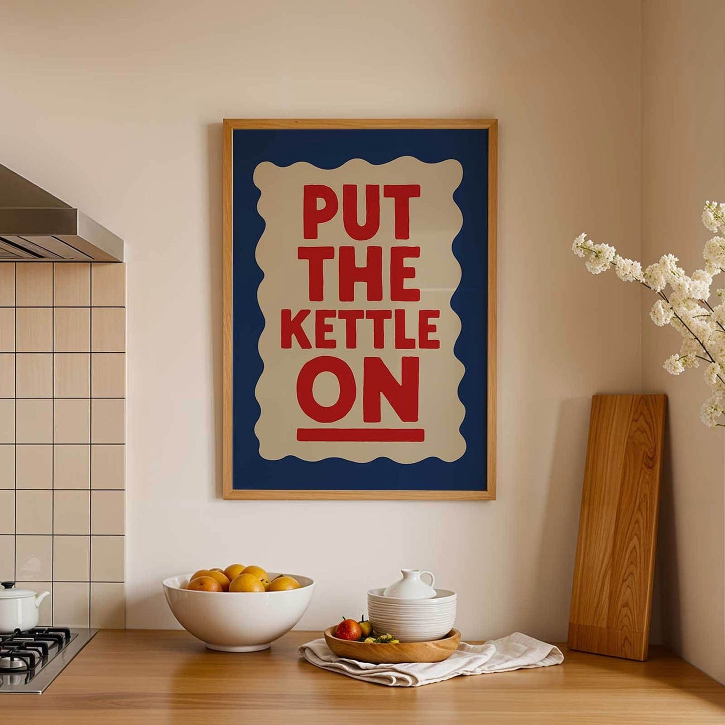 a kitchen counter with a framed poster that reads "PUT THE KETTLE ON" in bold red and blue letters. On the counter, there is a white bowl filled with oranges, a white teapot, and a wooden cutting board.