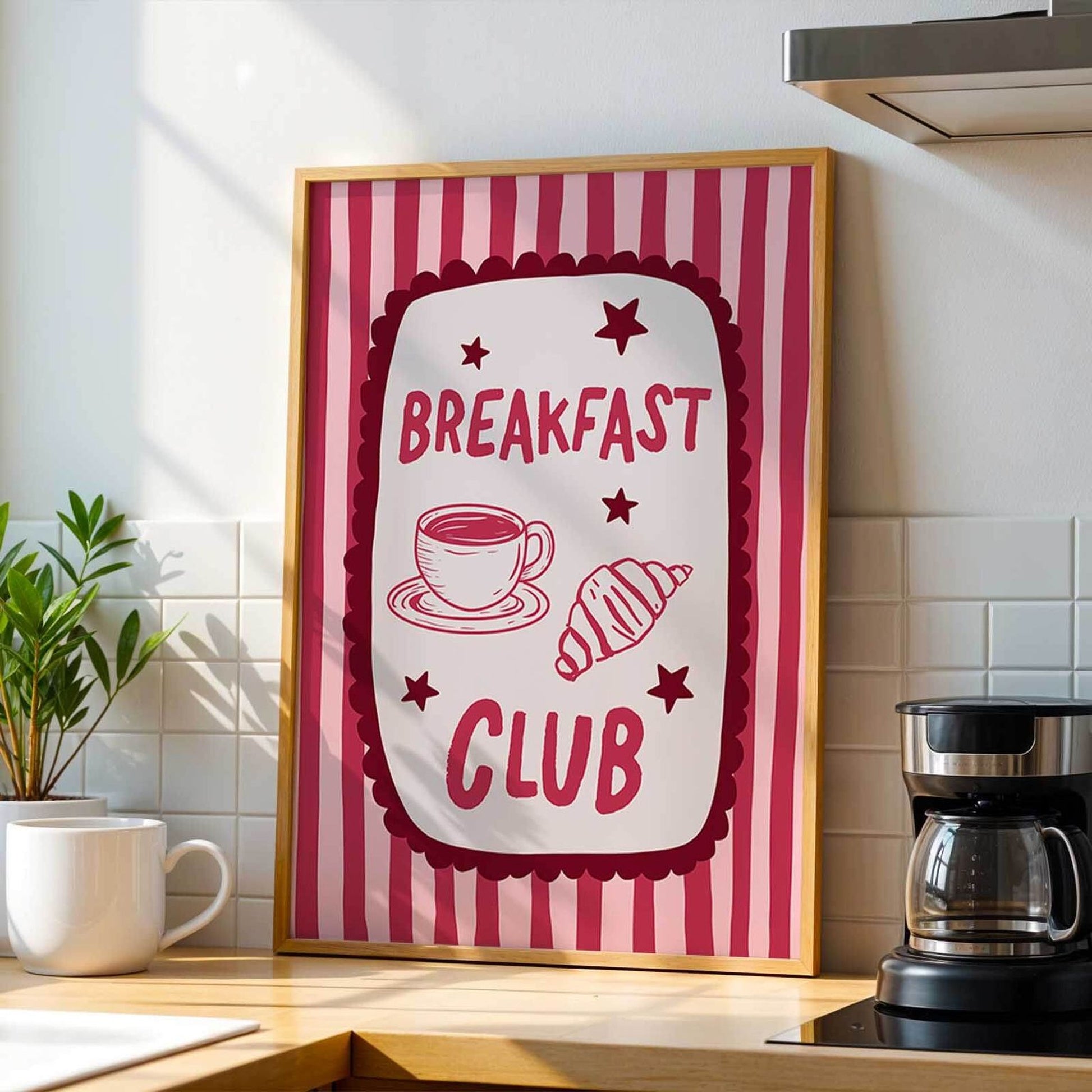 A framed poster with the text "BREAKFAST CLUB" and illustrations of a cup of coffee and a croissant, placed on a kitchen counter next to a coffee maker.