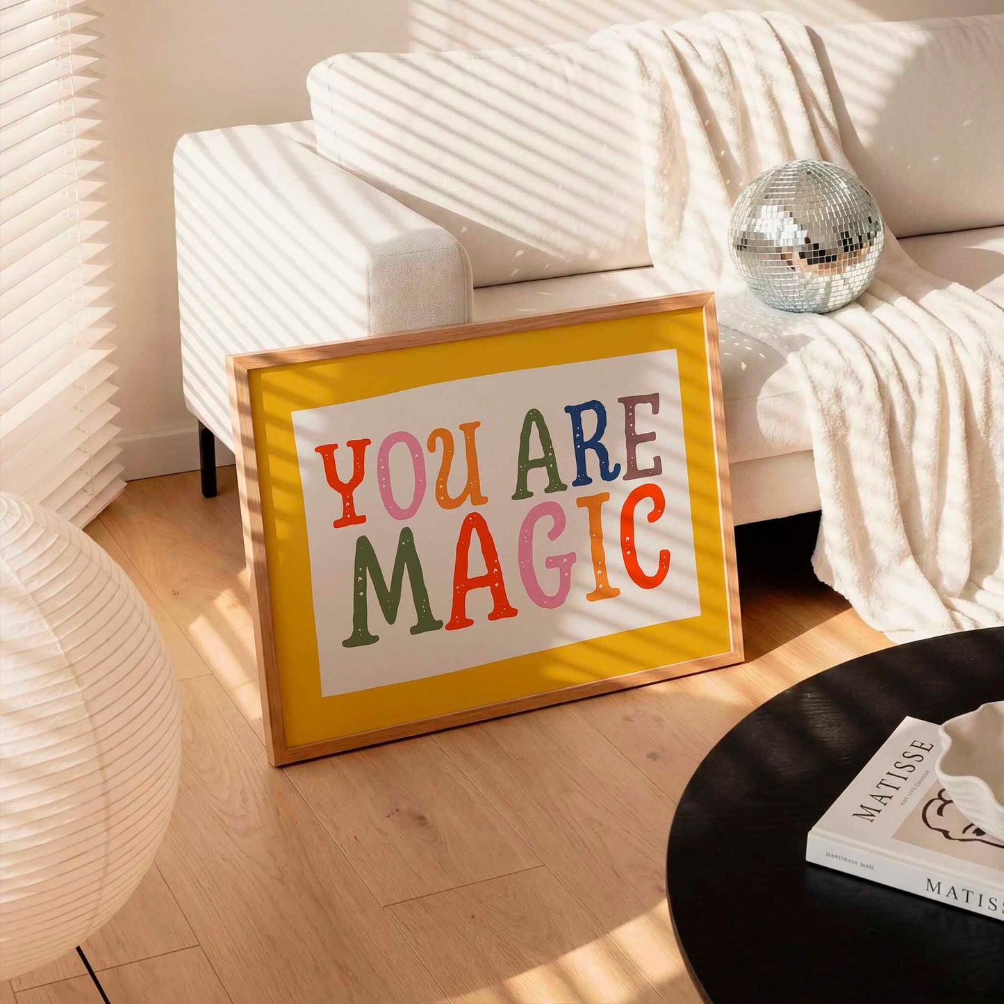 A framed poster with the text "YOU ARE MAGIC" is displayed on a wooden floor in a living room, with a white couch, a disco ball, and a lamp visible in the background.