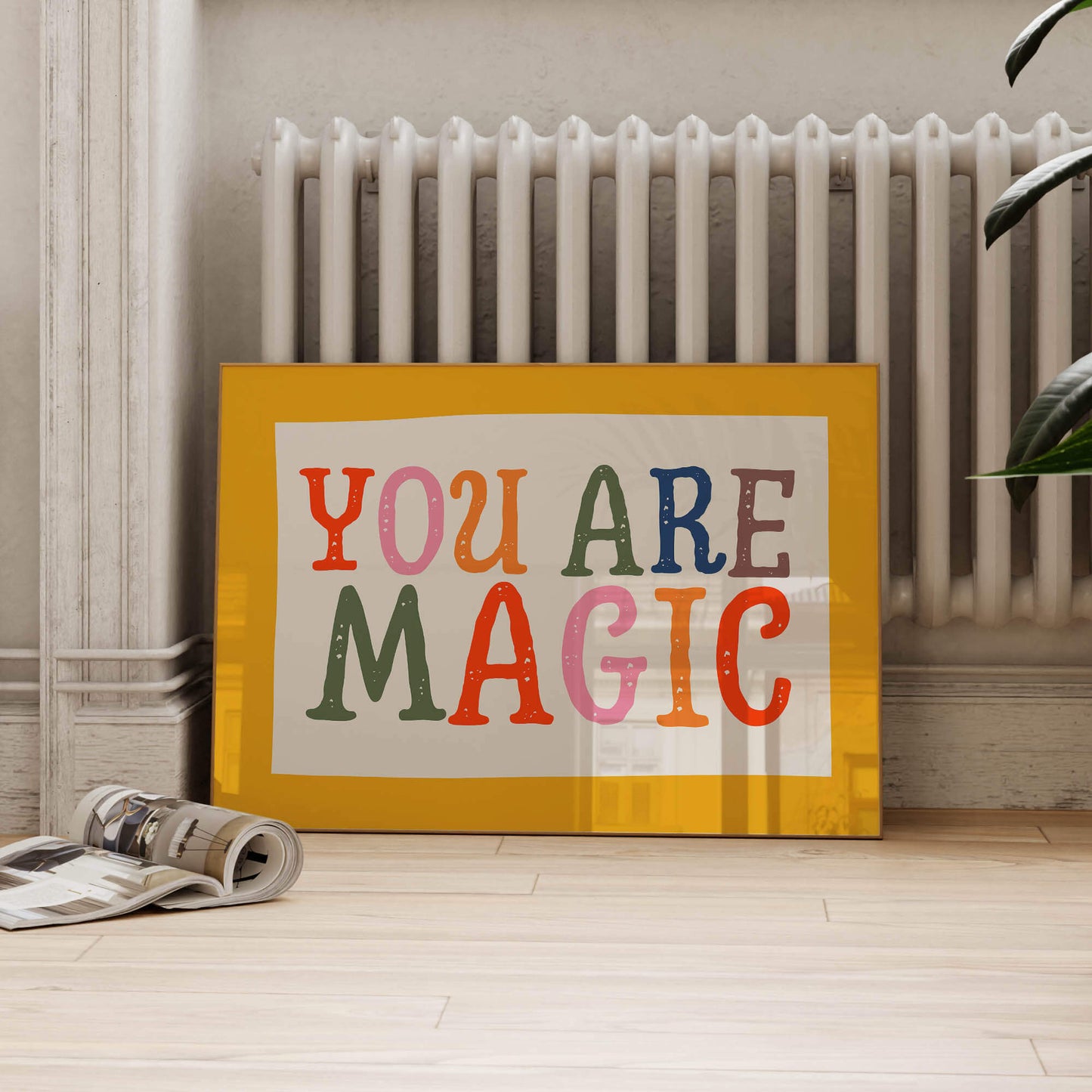 A framed poster with the text "YOU ARE MAGIC" is displayed on a wooden floor, with a white radiator visible in the background.