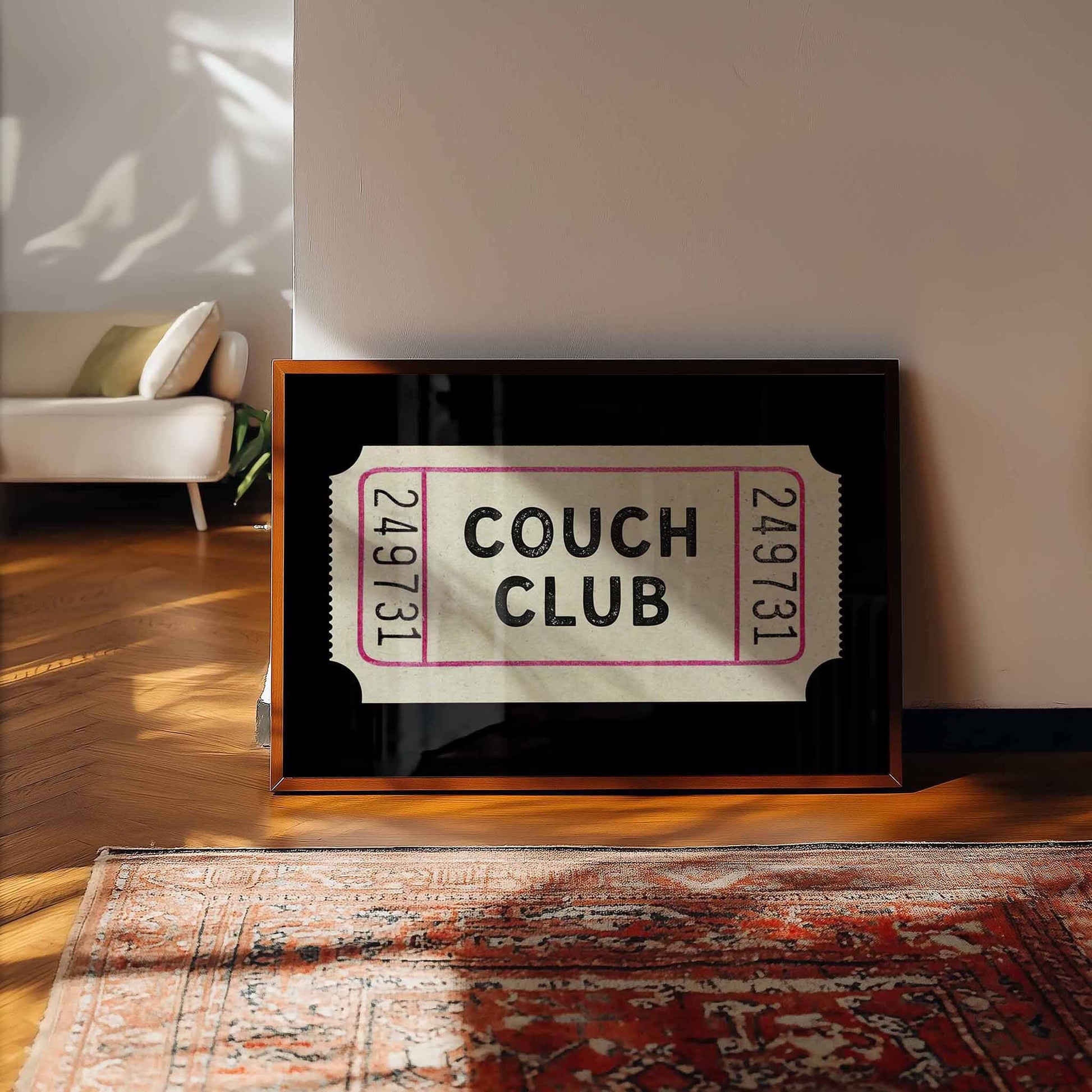 A framed ticket-like sign for the "Couch Club" is displayed on a wooden floor in a room with a white couch and a patterned rug.