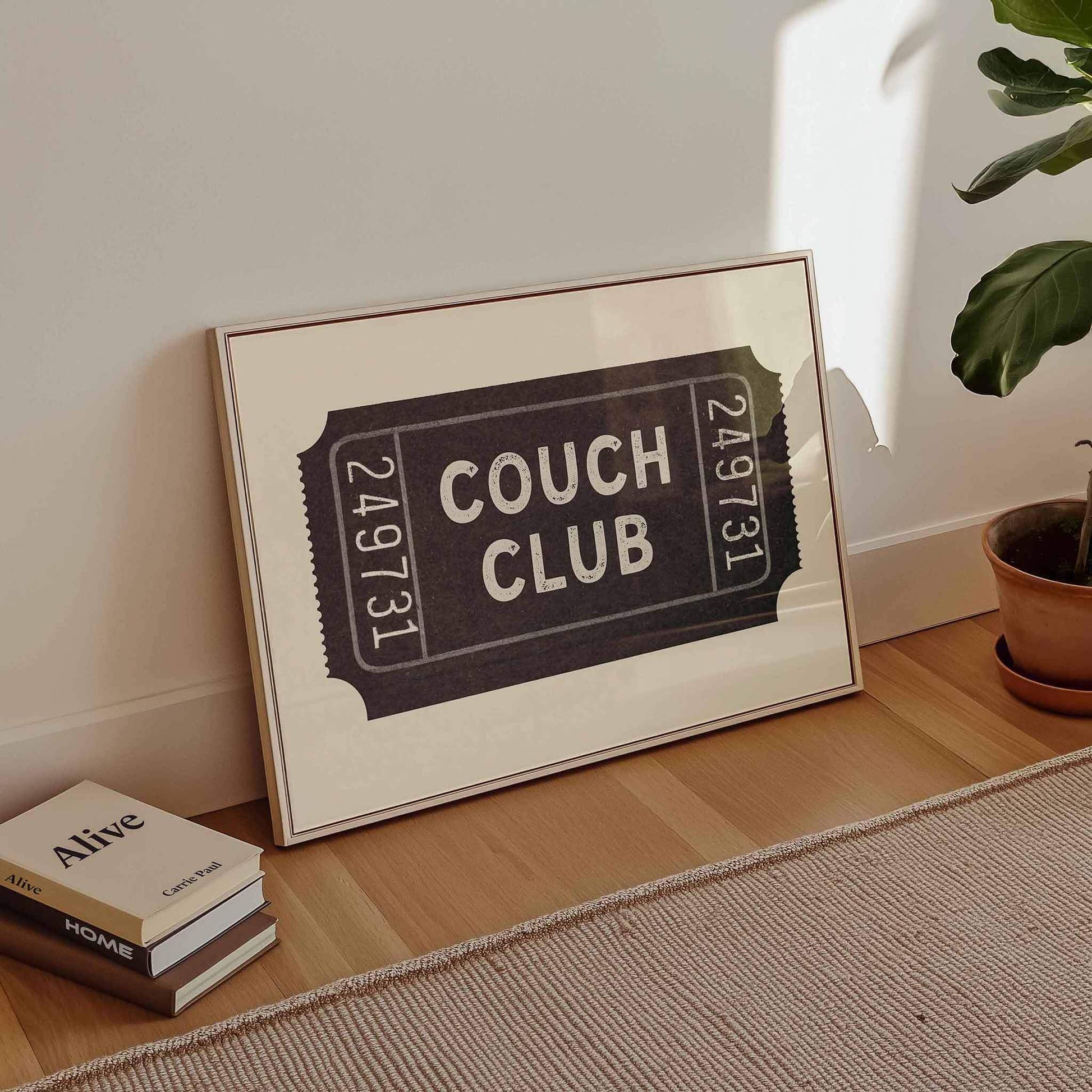 A framed poster with the text "Couch Club" is displayed on a wooden floor next to a stack of books and a potted plant.