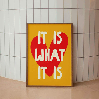 A framed poster with the text "IT IS WHAT IT IS" in bold letters, with a large red heart shape in the center.