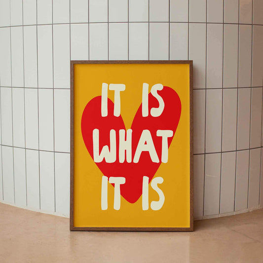 A framed poster with the text "IT IS WHAT IT IS" in bold letters, with a large red heart shape in the center.