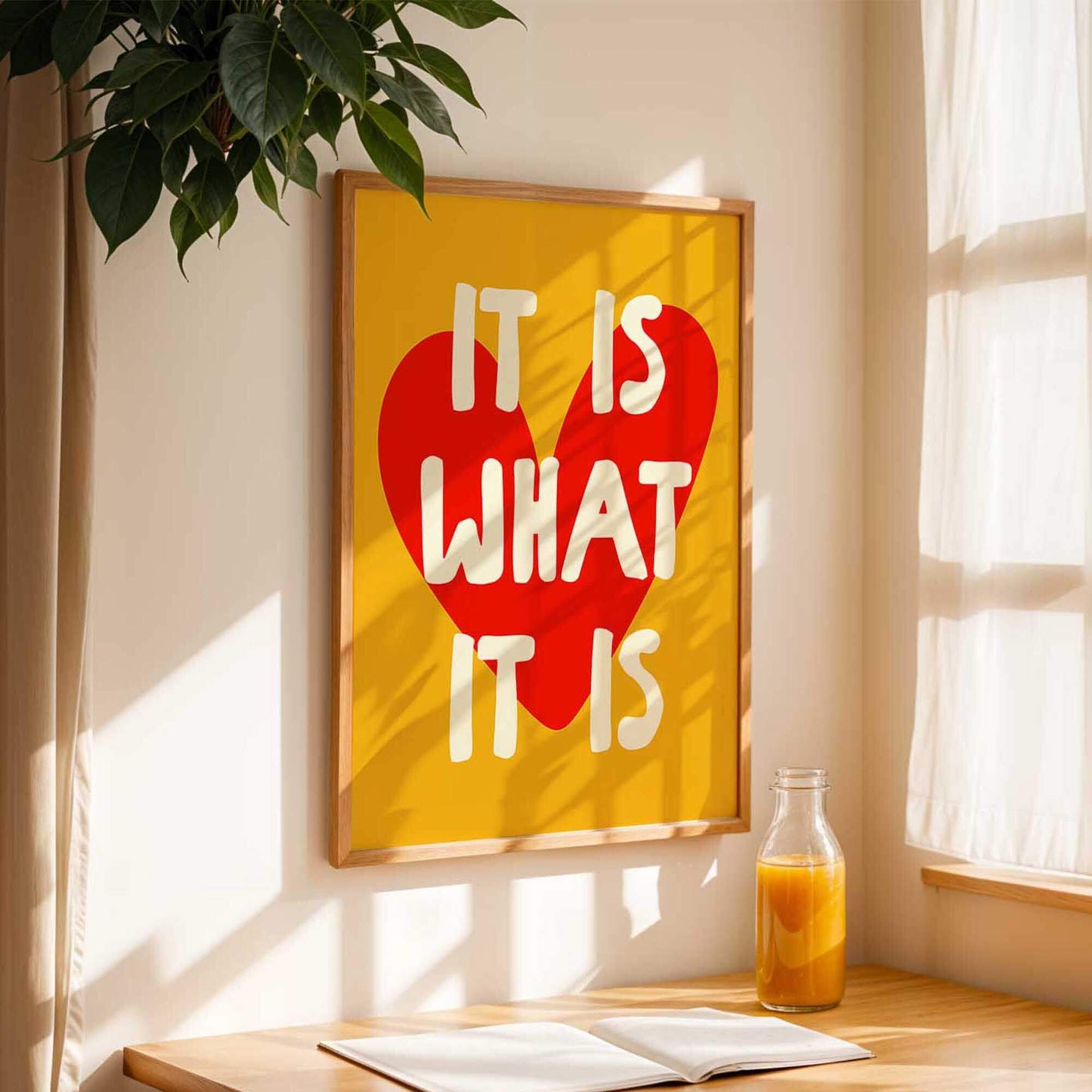 a framed poster with the text "IT IS WHAT IT IS" in bold letters, accompanied by a heart symbol, hanging on a wall next to a window. A bottle of orange juice is placed on a wooden table in front of the poster, and there is a book on the table as well.