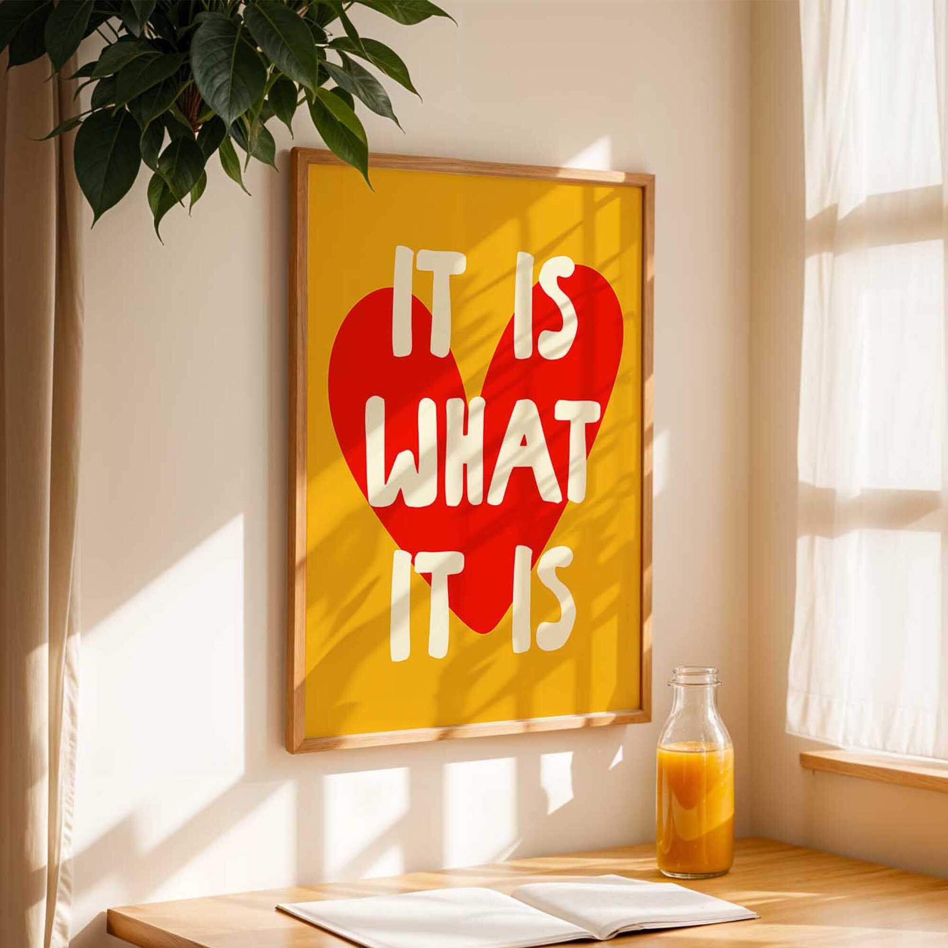 a framed poster with the text "IT IS WHAT IT IS" in bold letters, accompanied by a heart symbol, hanging on a wall next to a window. A bottle of orange juice is placed on a wooden table in front of the poster, and there is a book on the table as well.