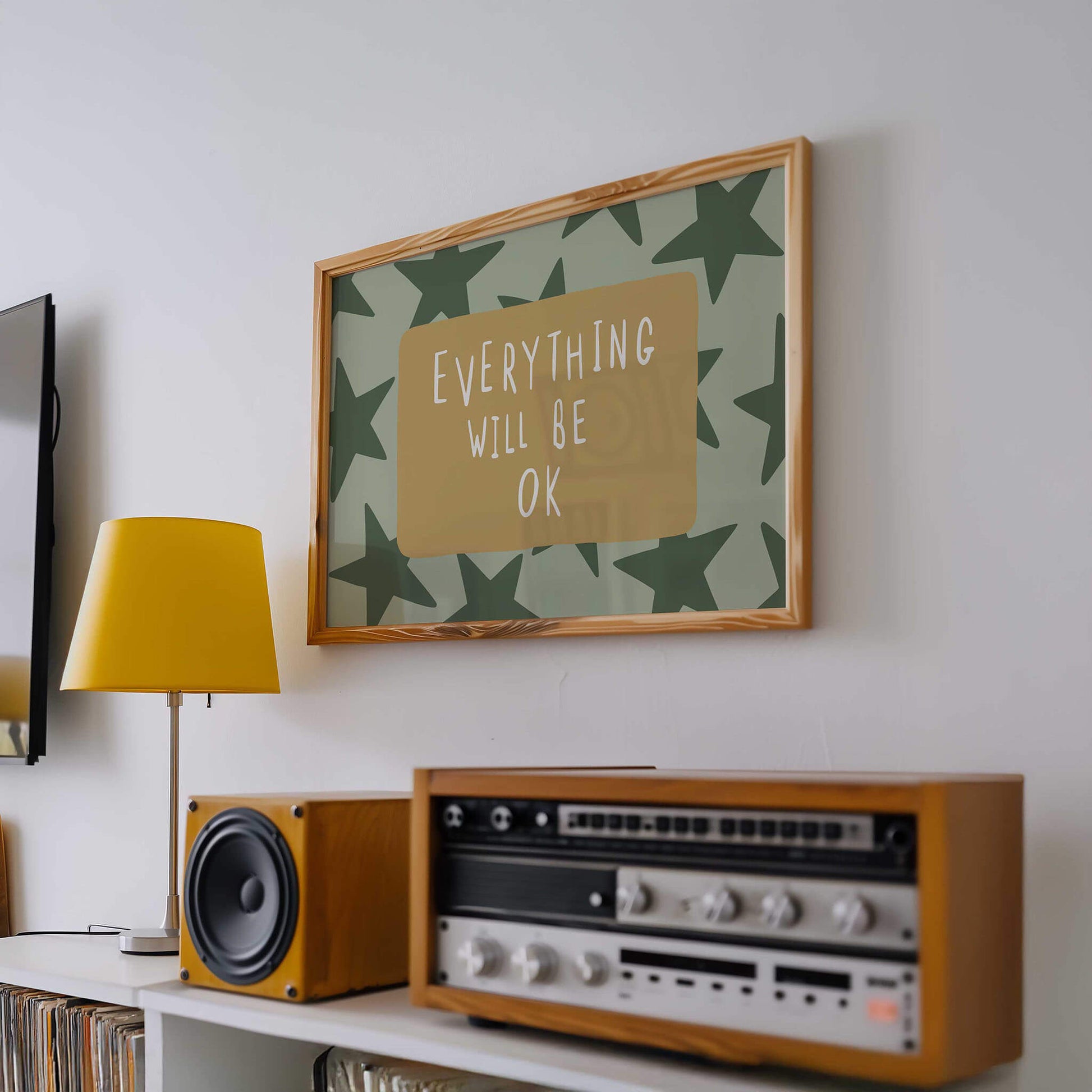 a framed poster with the text "EVERYTHING WILL BE OK" on a wall, accompanied by a wooden radio and a yellow lamp on a shelf below it.