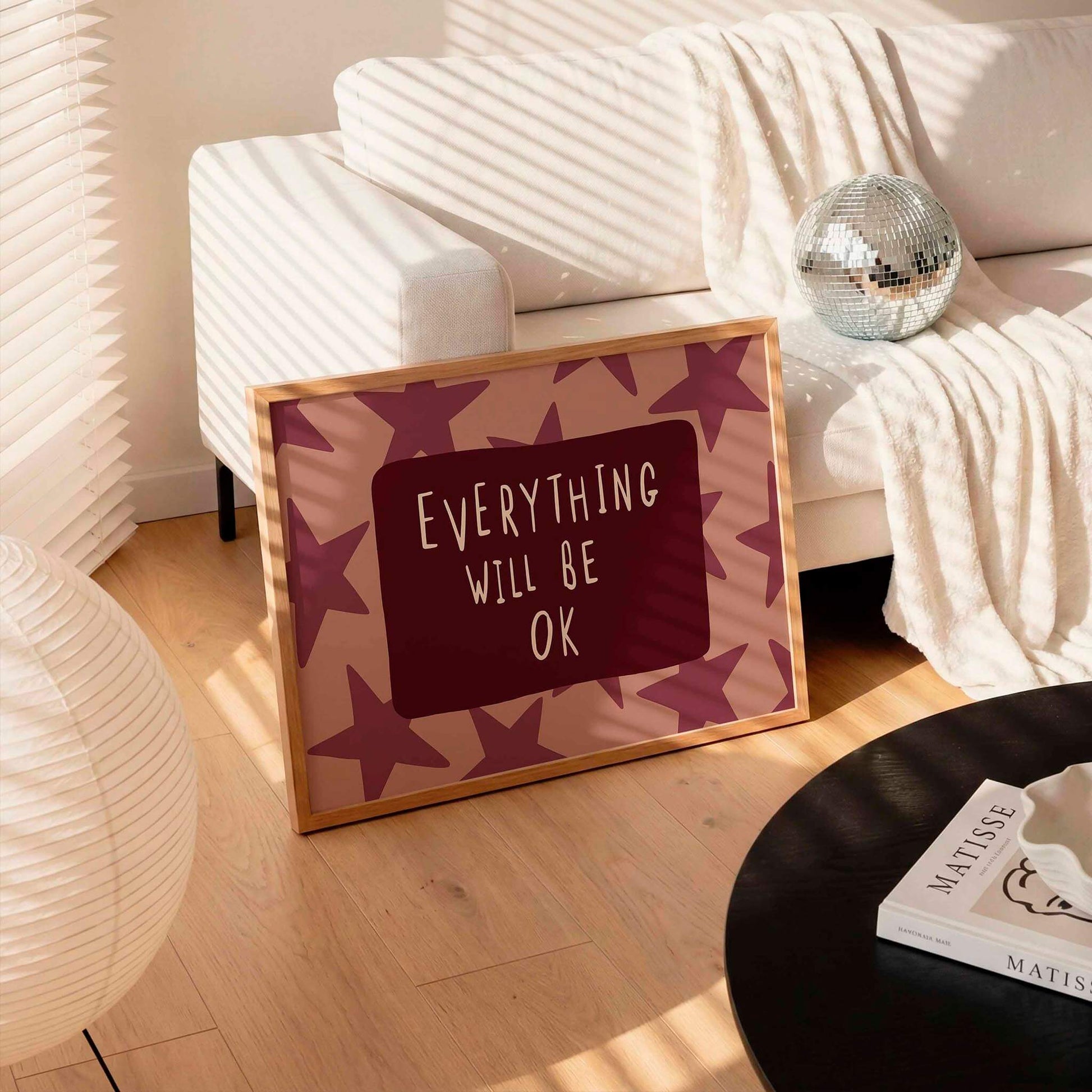 a framed sign with the text "EVERYTHING WILL BE OK" on a wooden floor, surrounded by a white couch, a silver disco ball, and a lamp.