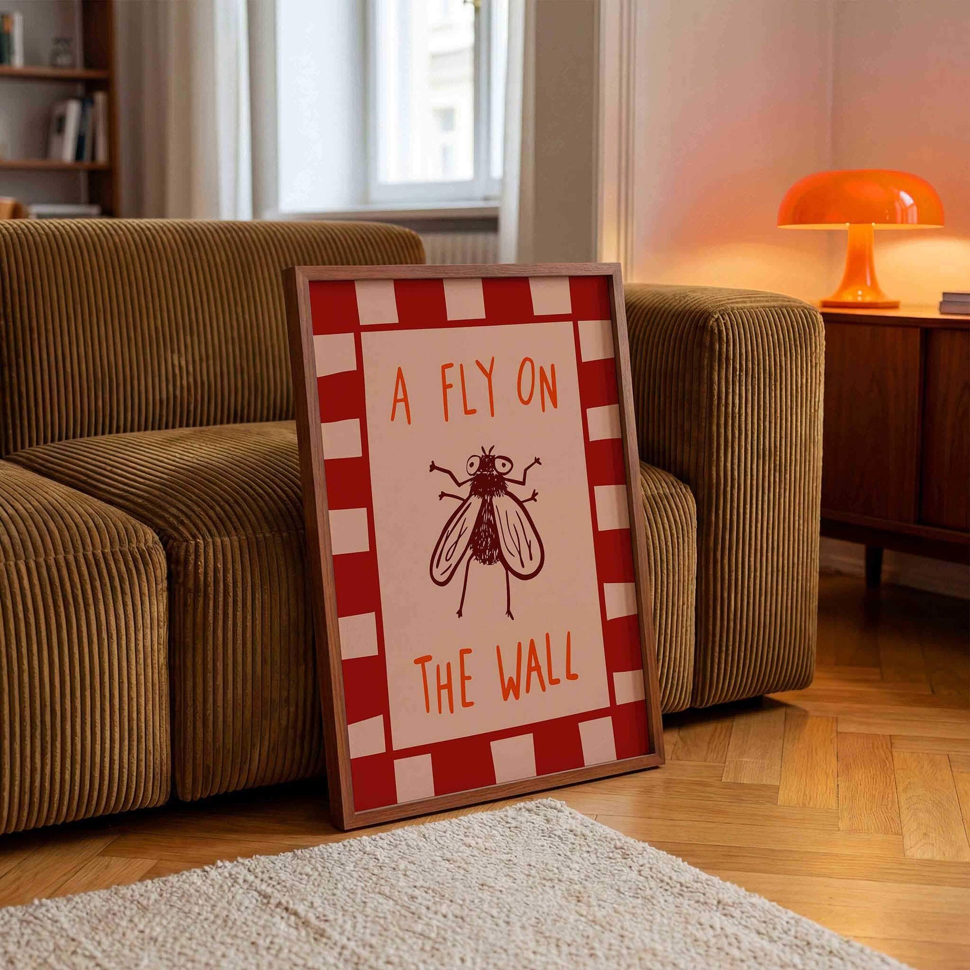 A framed poster with the text "A FLY ON THE WALL" is displayed on a wooden floor in a living room, with a couch and a lamp visible in the background.