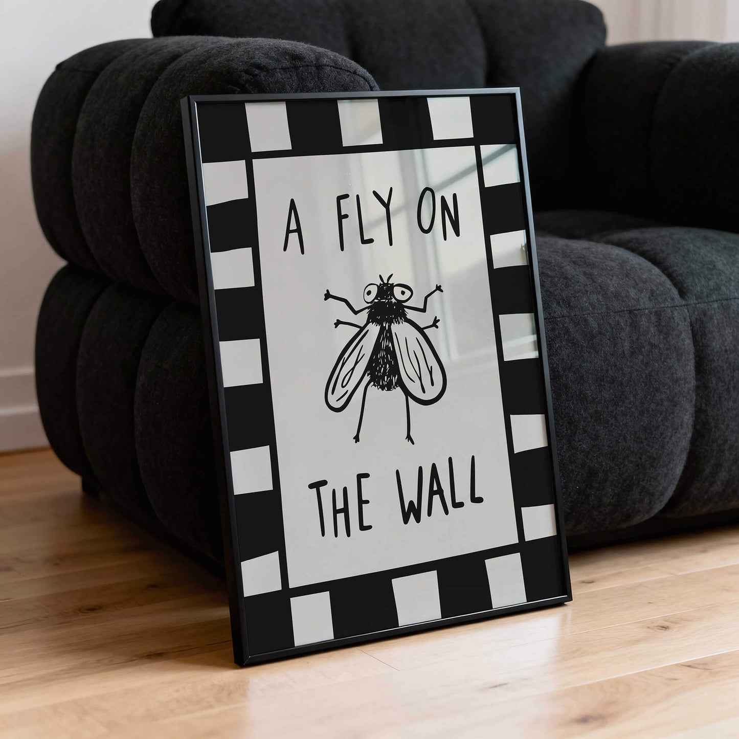 A framed poster with the text "A FLY ON THE WALL" and a black and white illustration of a fly is placed on a black couch.