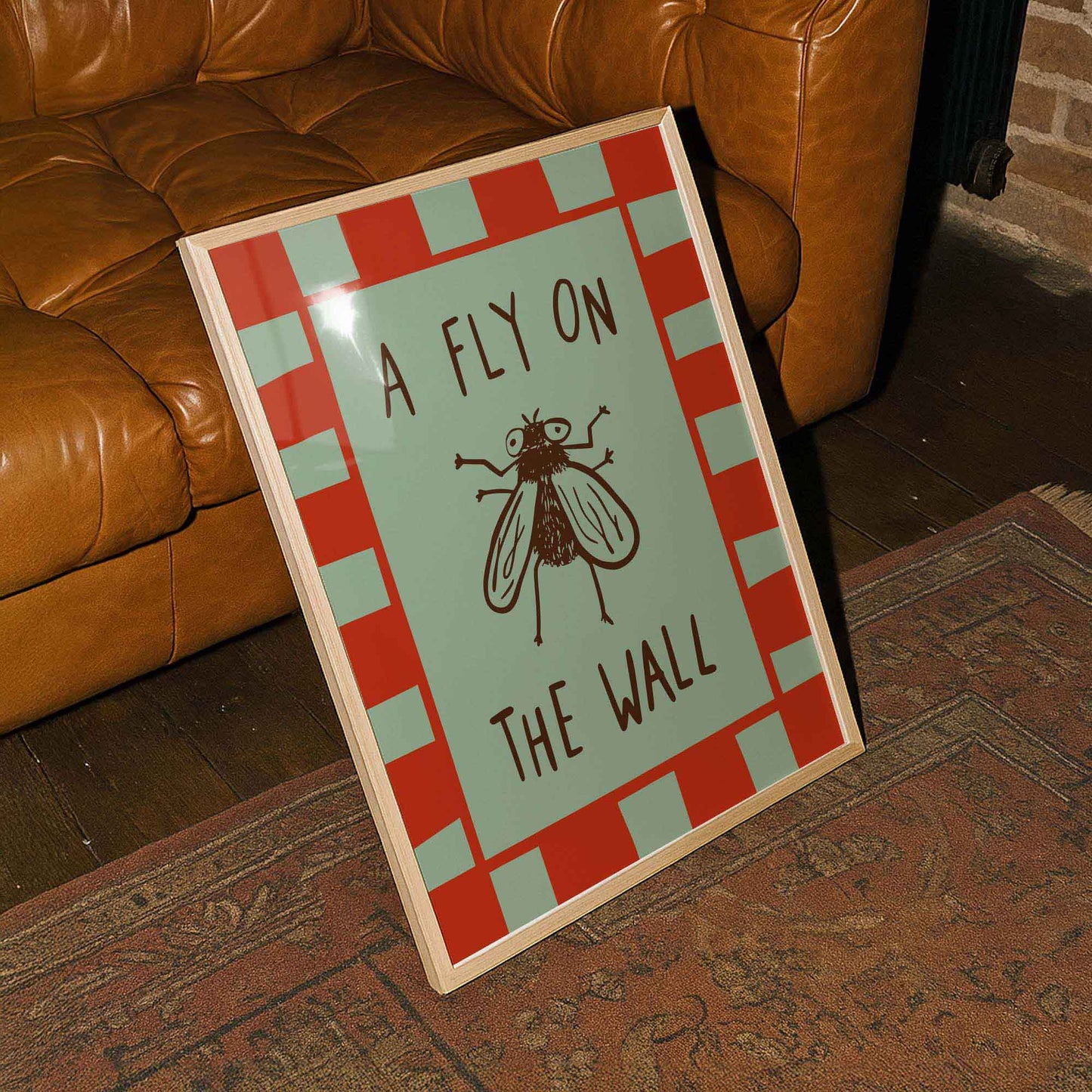A framed poster with the text "A FLY ON THE WALL" and an illustration of a fly is placed on a rug next to a brown leather couch.