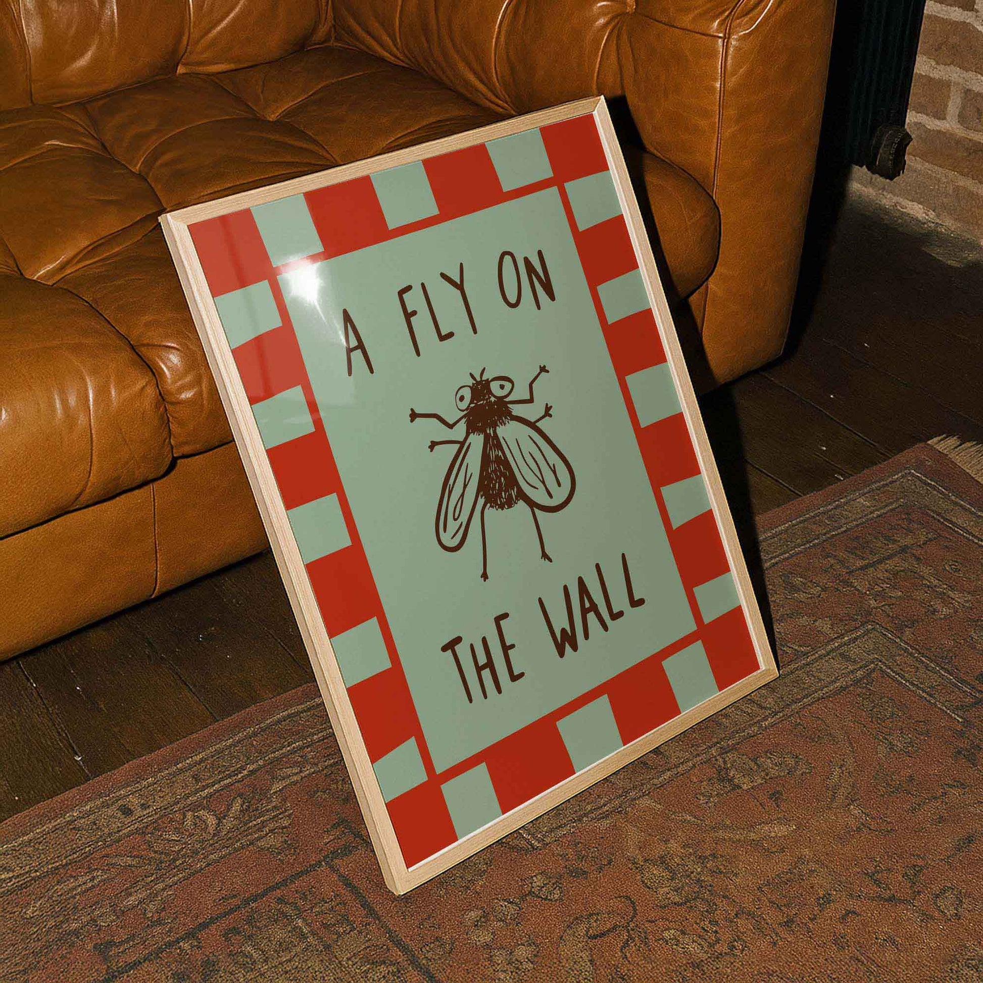 A framed poster with the text "A FLY ON THE WALL" and an illustration of a fly is placed on a rug next to a brown leather couch.