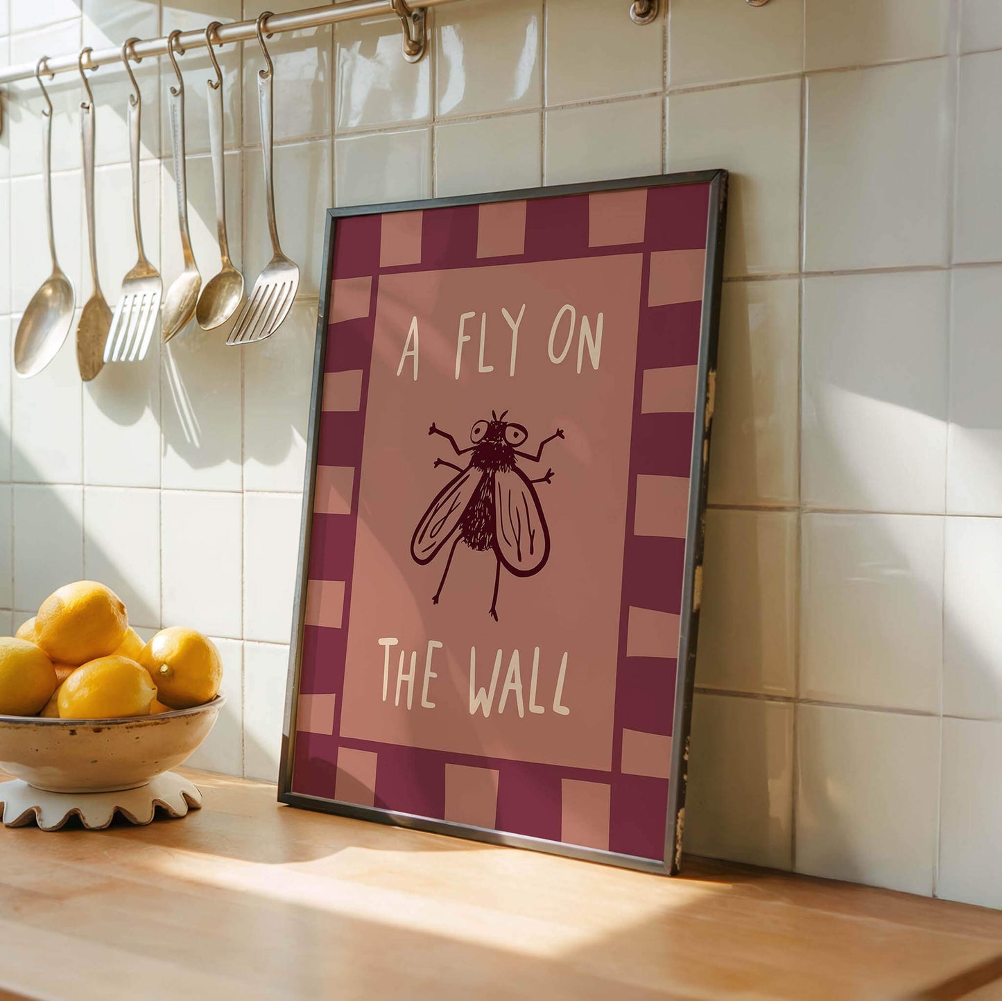 A framed poster with the text "A FLY ON THE WALL" is displayed on a kitchen counter, with a bowl of lemons and a set of spoons hanging nearby.