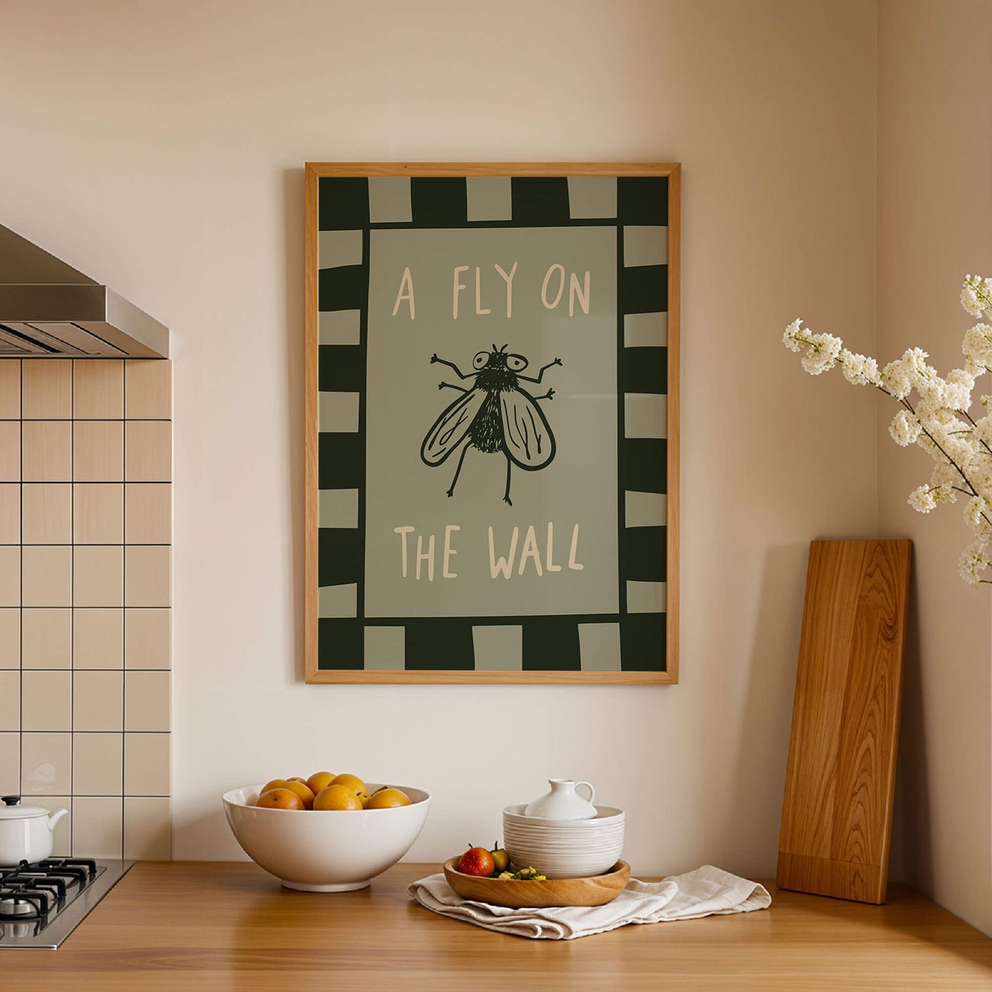 a kitchen counter with a framed poster of a fly on the wall above it, a bowl of fruit, and a wooden cutting board.