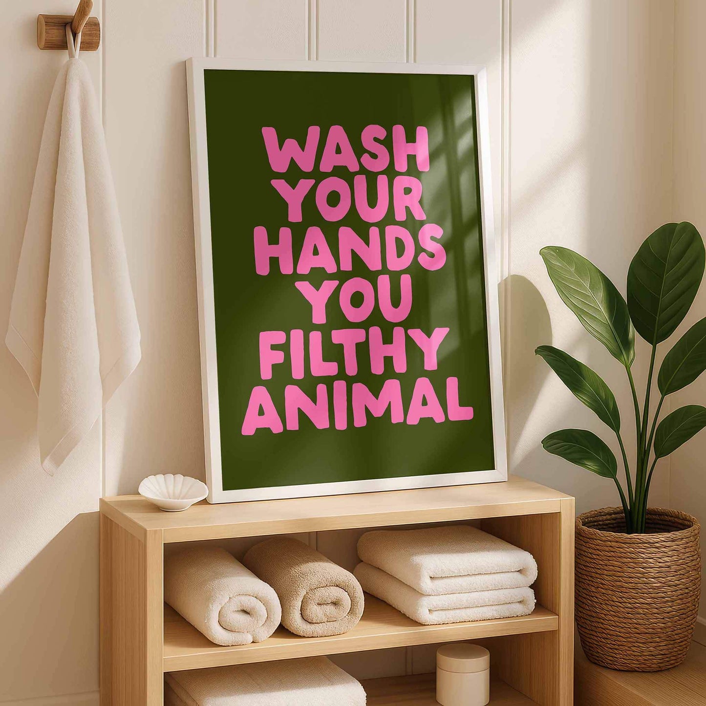 a framed poster with the text "WASH YOUR HANDS YOU FILthy ANIMAL" in pink letters, placed on a wooden shelf next to a potted plant.