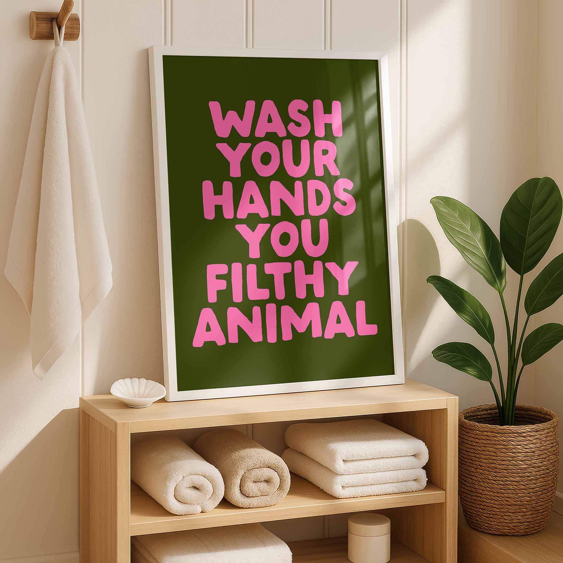 a framed poster with the text "WASH YOUR HANDS YOU FILthy ANIMAL" in pink letters, placed on a wooden shelf next to a potted plant.