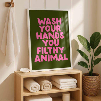 a framed poster with the text "WASH YOUR HANDS YOU FILthy ANIMAL" in pink letters, placed on a wooden shelf next to a potted plant.