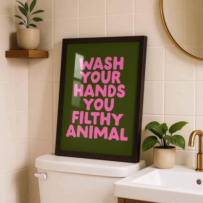 a framed poster with the text "WASH YOUR HANDS YOU FILthy ANIMAL" displayed on a bathroom wall.
