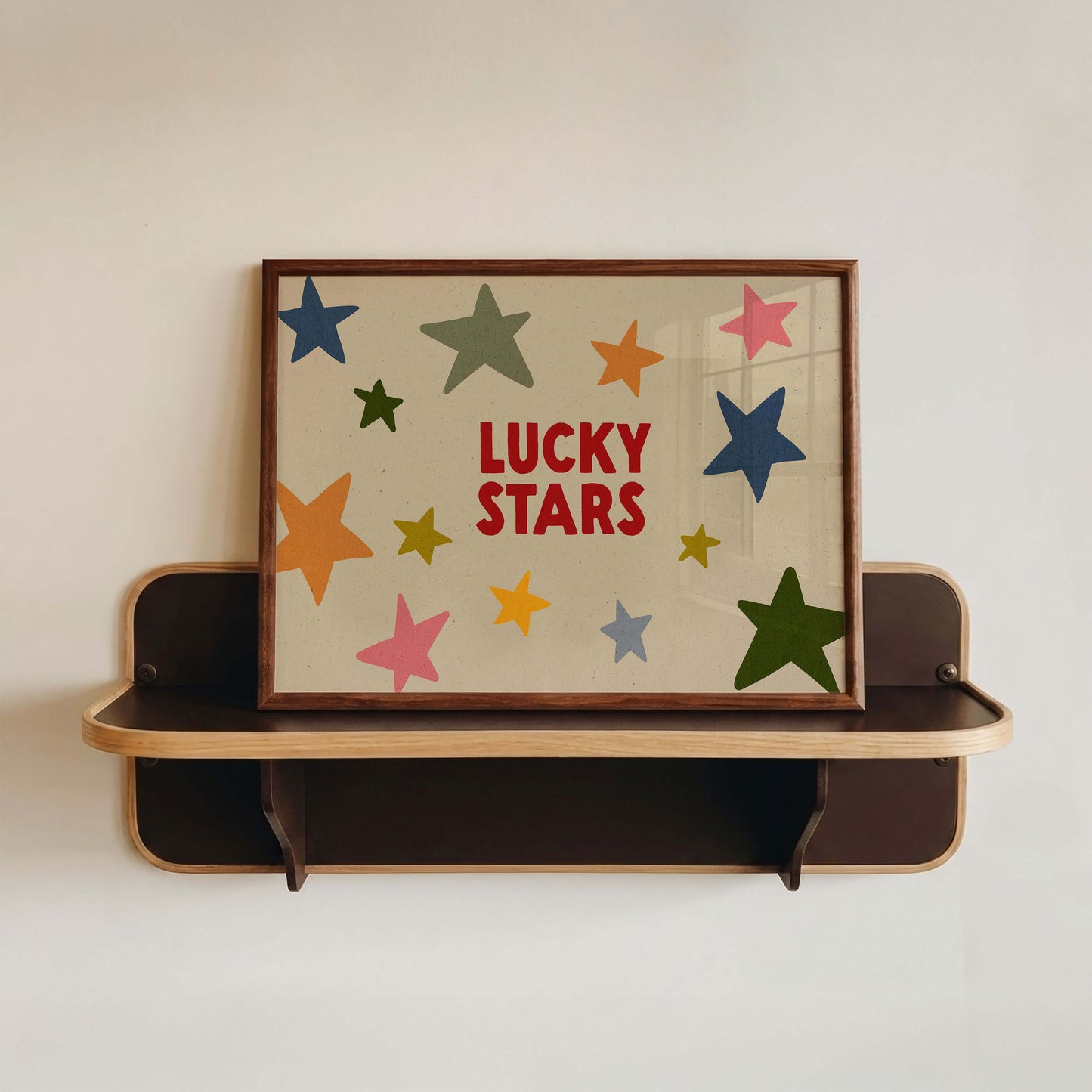 A framed wall art print with the words 'LUCKY STARS' in red, surrounded by colorful star shapes, displayed on a dark wooden floating shelf against a plain white wall.