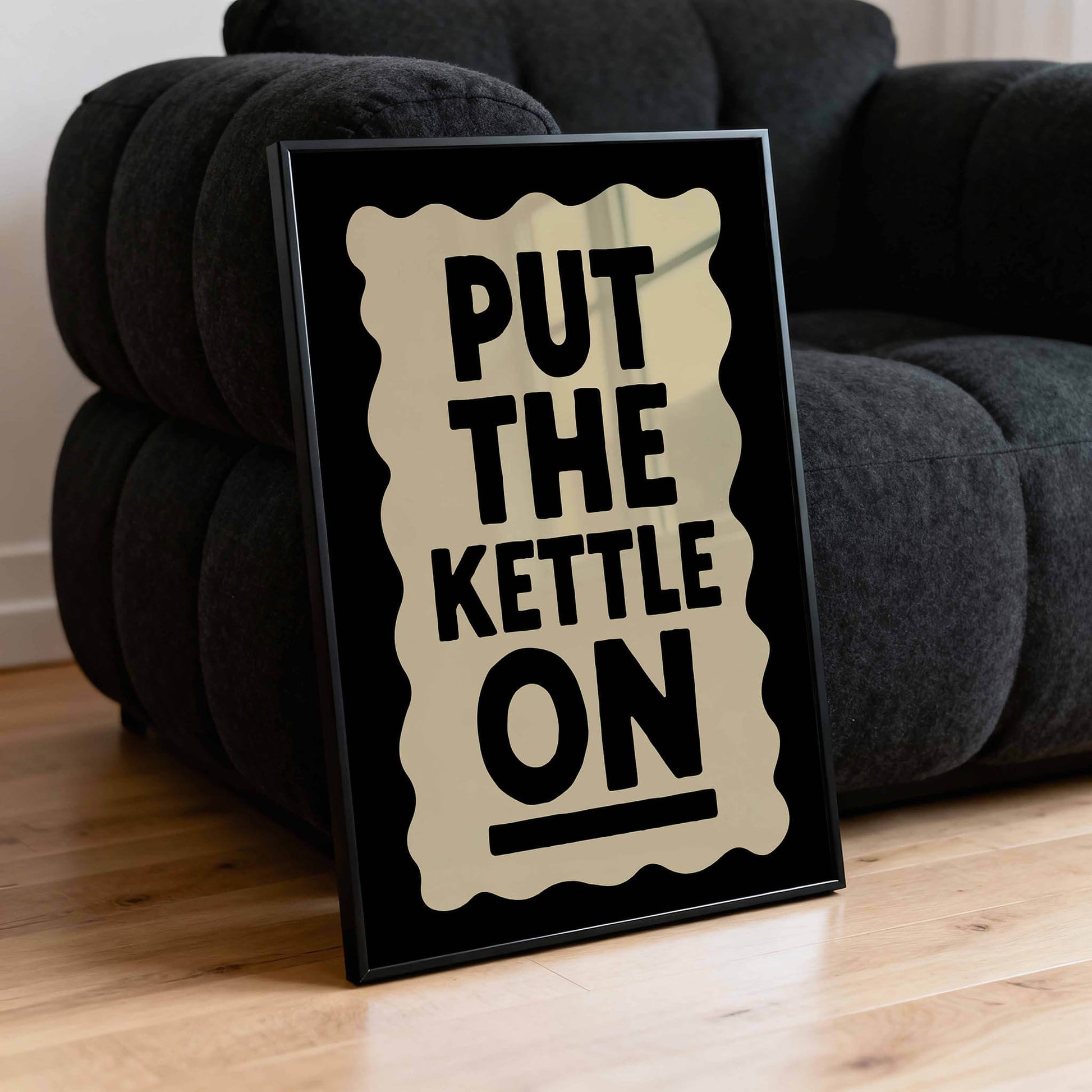 A framed poster with the text "PUT THE KETTLE ON" is placed on a black couch.