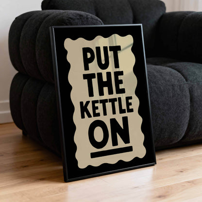 A framed poster with the text "PUT THE KETTLE ON" is placed on a black couch.