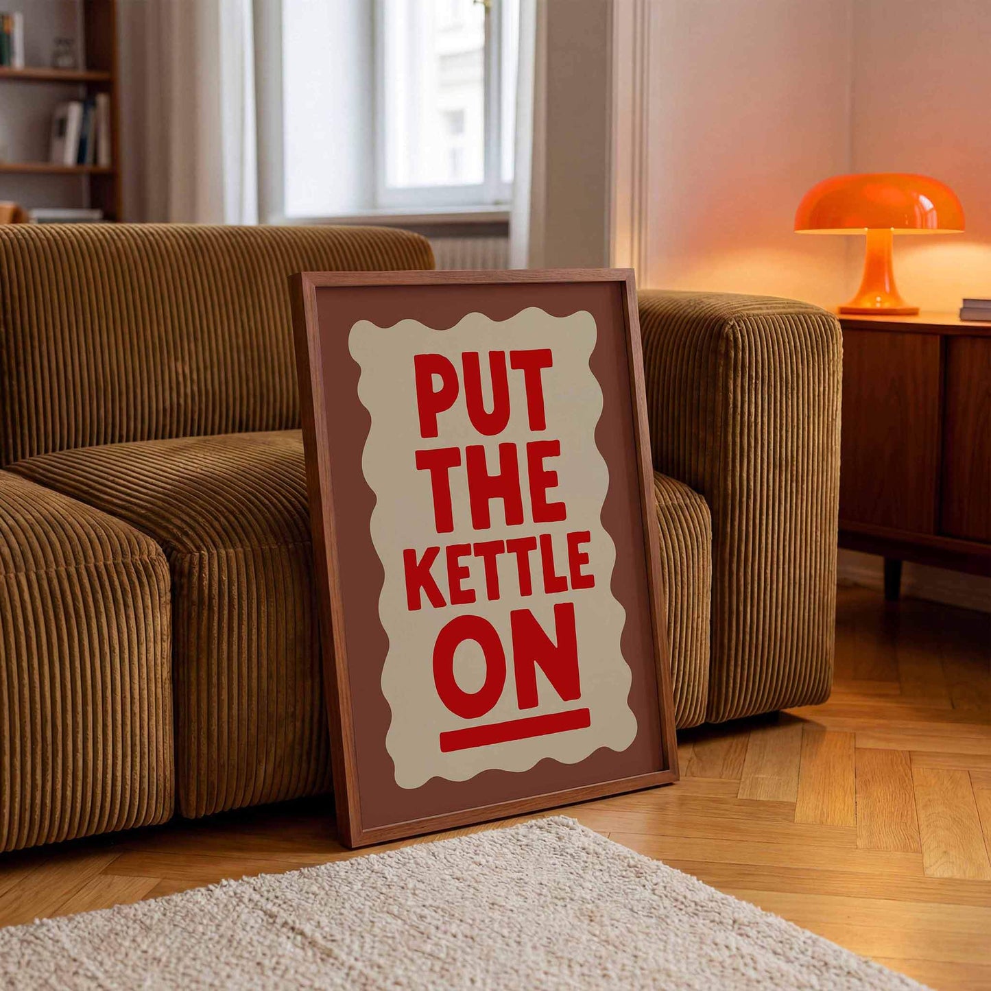 A framed poster with the text "PUT THE KETTLE ON" is displayed on a wooden floor in a living room.