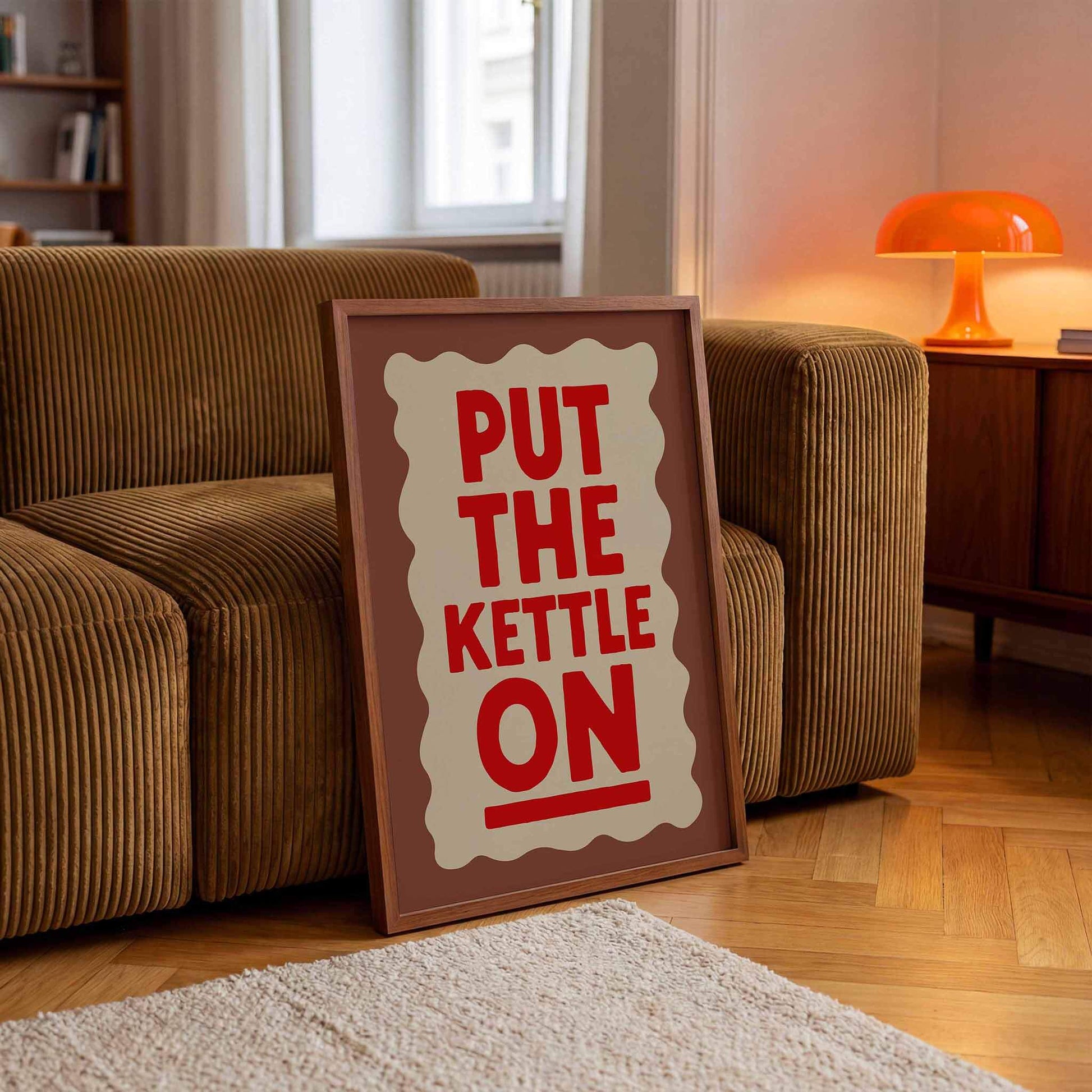 A framed poster with the text "PUT THE KETTLE ON" is displayed on a wooden floor in a living room.