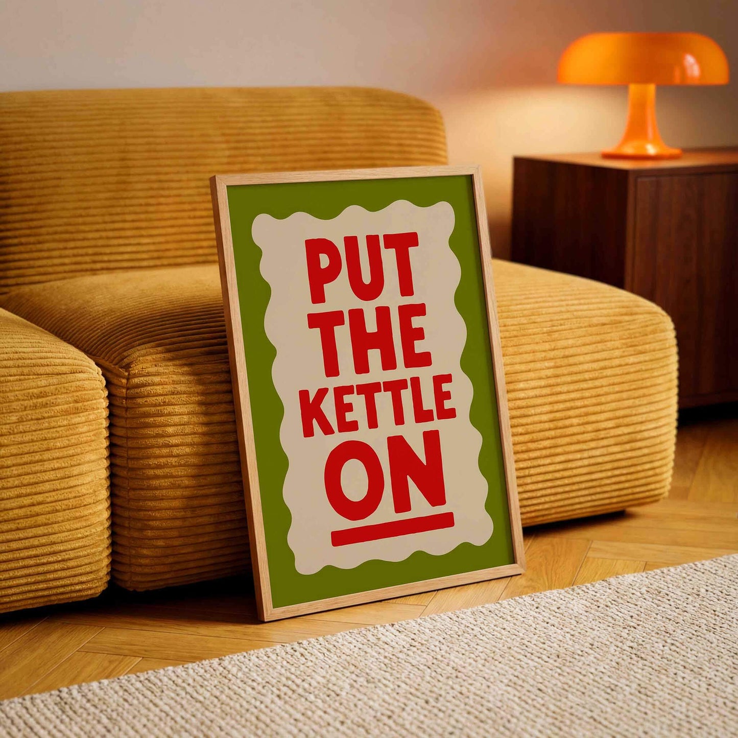A framed poster with the text "PUT THE KETTLE ON" is displayed on a wooden floor in a living room, with a yellow couch and a lamp in the background.