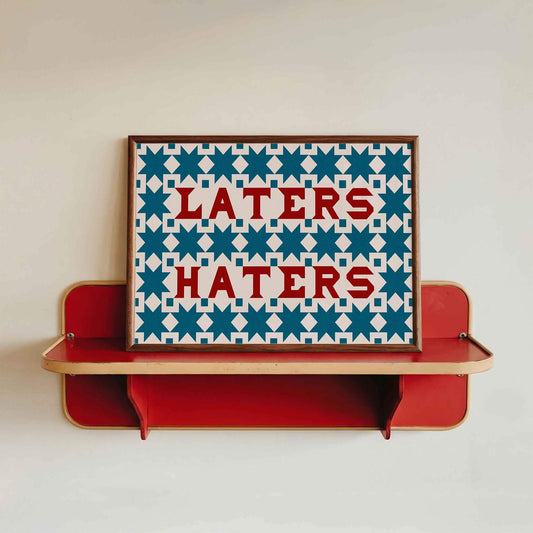 A retro and boho statement wall art print in red and blue made of quilting squares and the words LATERS HATERS. Displayed in a horizontal wooden frame sitting on a retro red shelf.