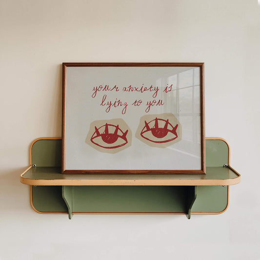 a framed artwork with a message about anxiety and lying, displayed on a shelf against a white wall.