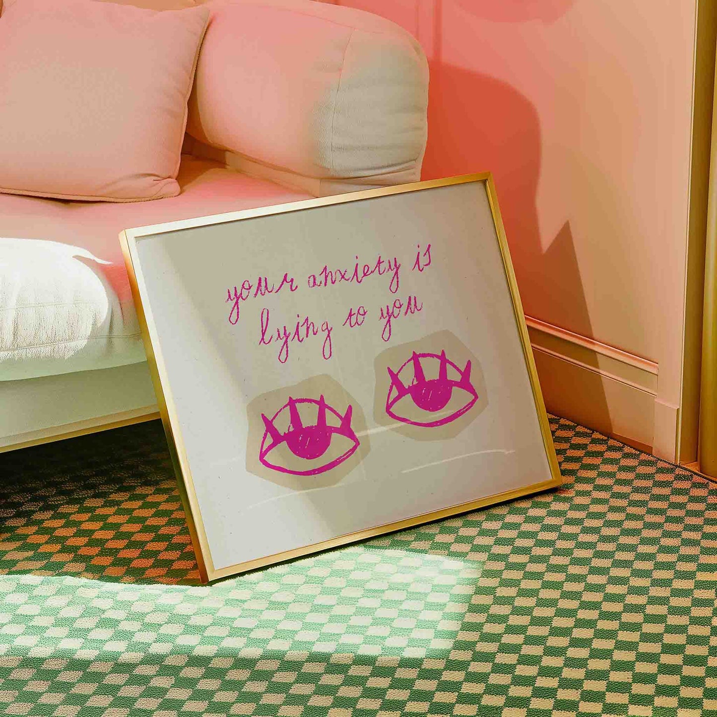 a framed piece of art with a pink and white color scheme, placed on a green and white checkered floor. The art piece has a message that reads "your anxiety is lying to you" and includes two pink eye-like symbols.