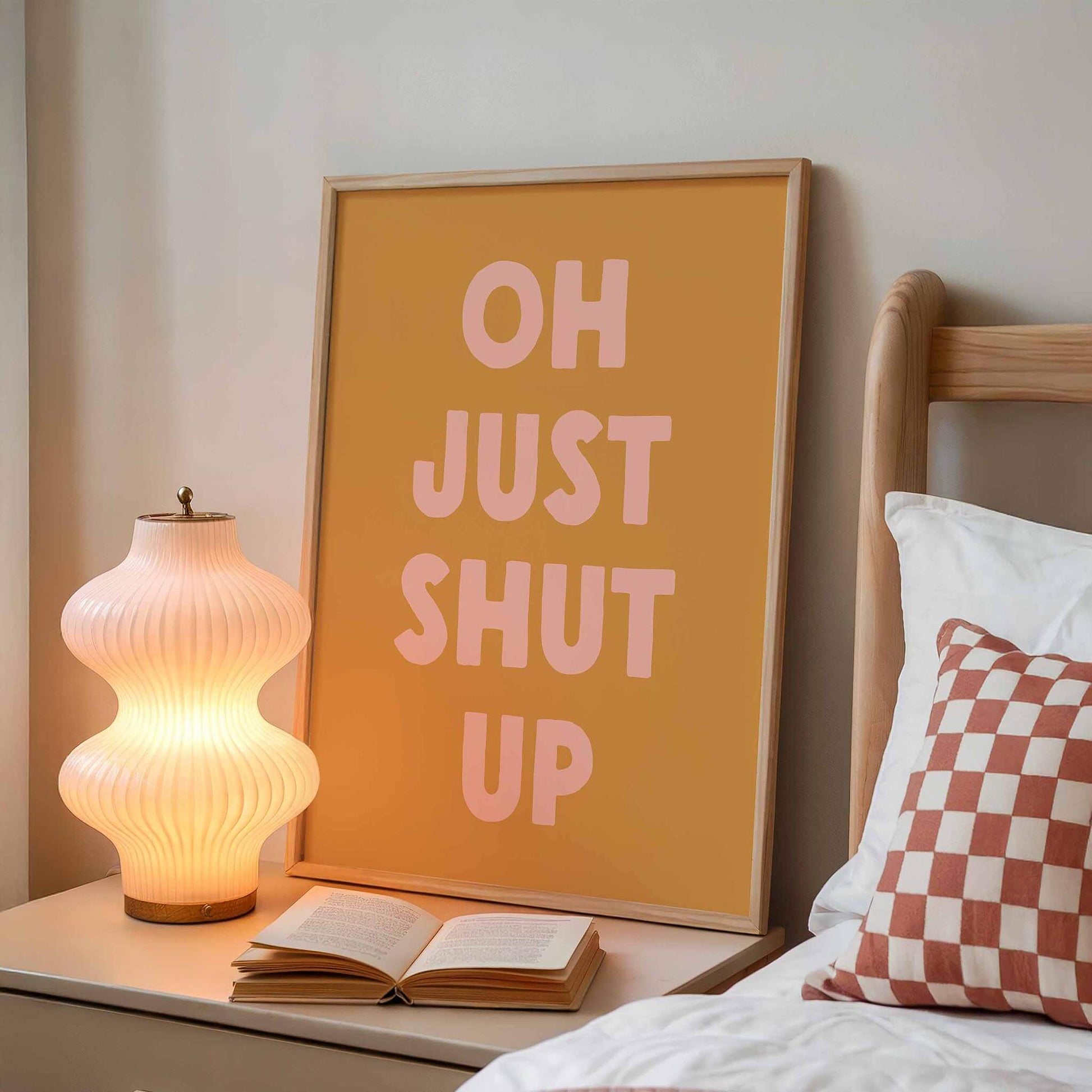 a bedside table with a lamp, an open book, and a framed poster with the text "OH JUST SHUT UP" on it.