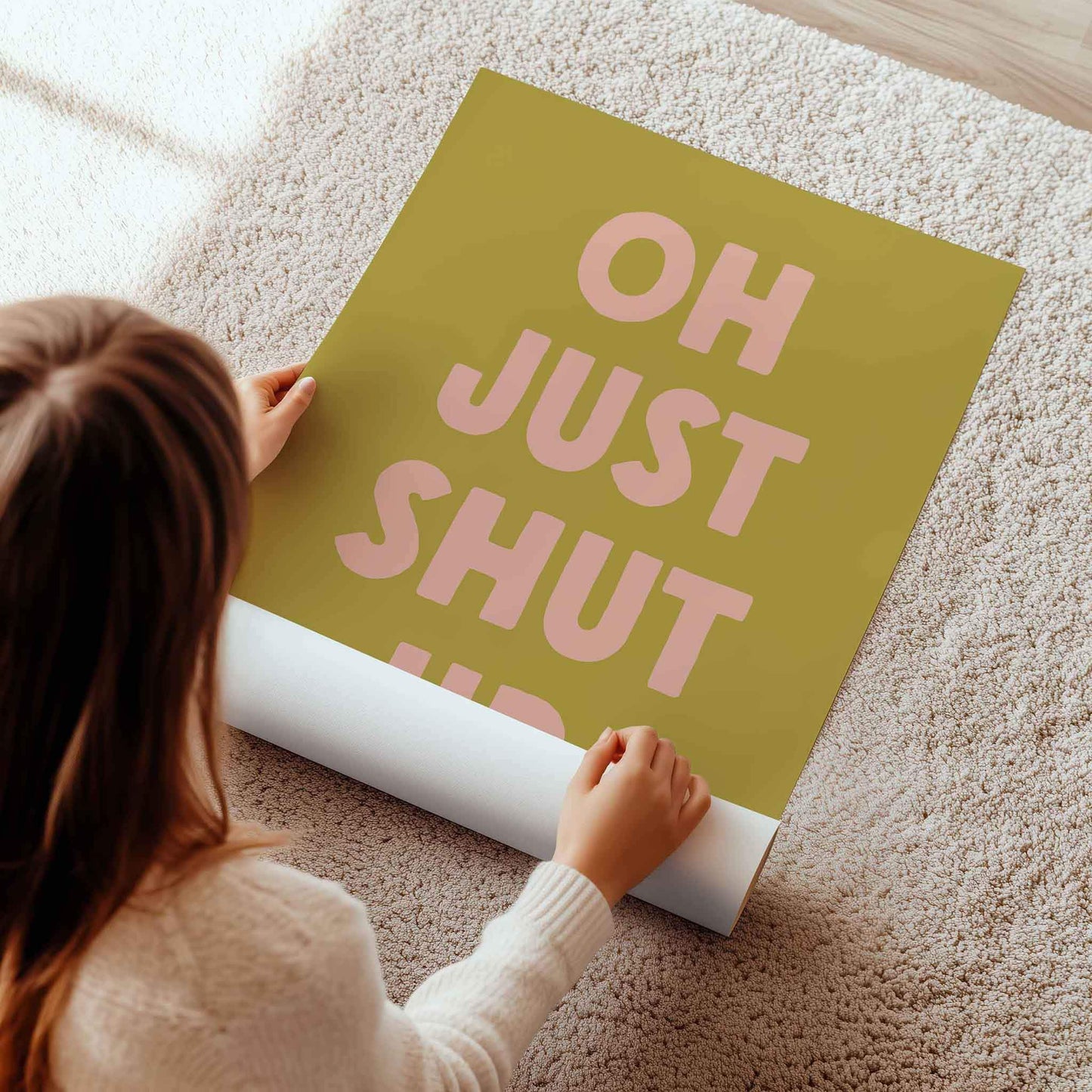 A person is laying on the floor and holding a roll of paper, which appears to be a poster or a sign with the text "OH JUST SHUT UP" written on it.