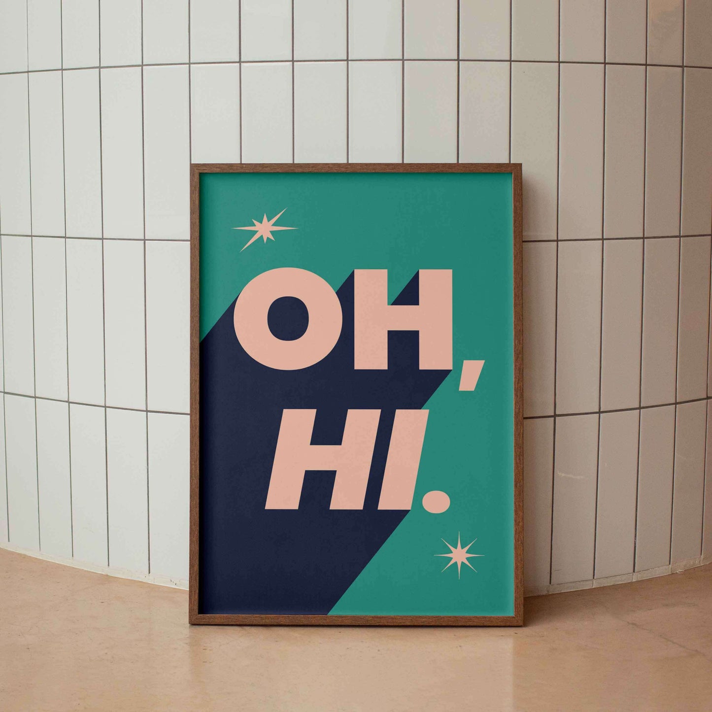 A framed typographic art print leaning against a white tiled wall on a beige floor. The print features the words “OH, HI.