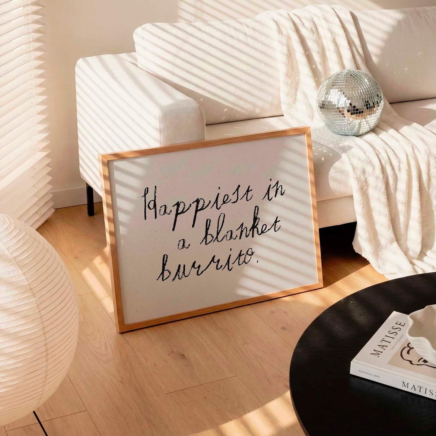 A framed sign with the text "happiest in a blanket burrito" is displayed in a living room setting, with a white couch, a lamp, and a disco ball visible in the background.
