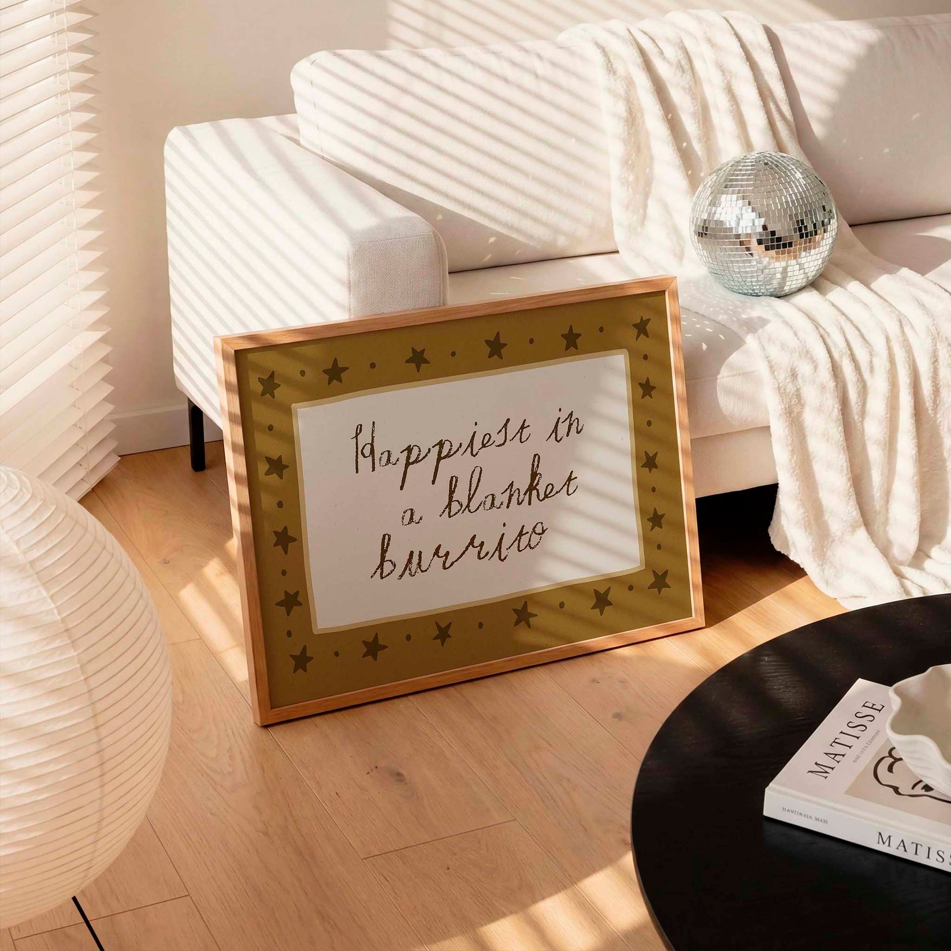 A framed sign with the text "Happiest in a blanket burrito" is displayed on a wooden floor in a living room, with a white couch and a disco ball in the background.