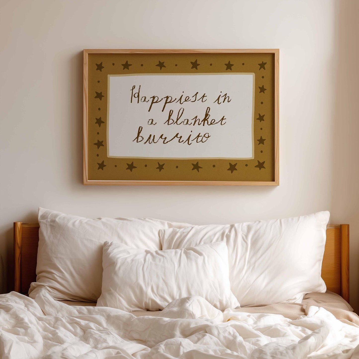 a cozy bedroom scene with a bed covered in white sheets and pillows. Above the bed, there is a framed wall art with the text "Happiest in a blanket burrito" written in a decorative font.