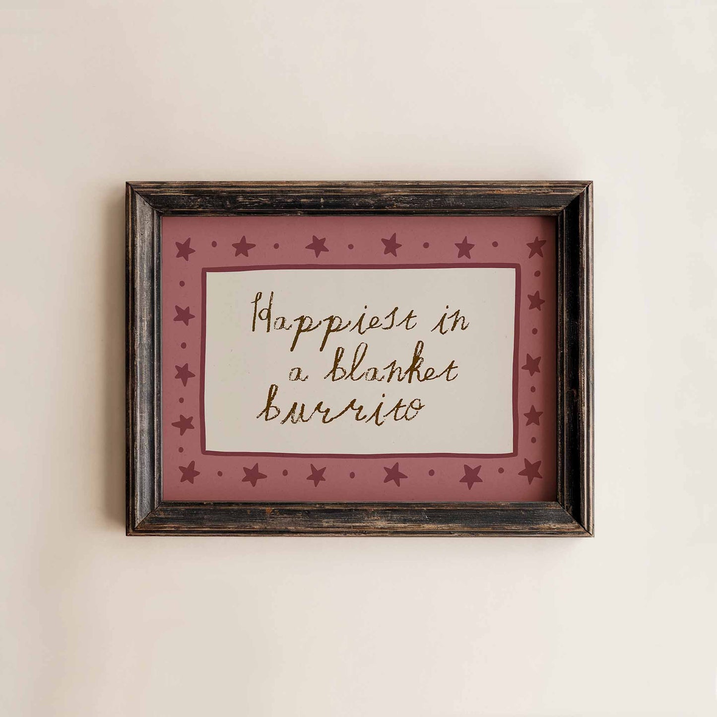 A framed picture with a pink border and stars, containing the handwritten text "Happiest in a blanket burrito".