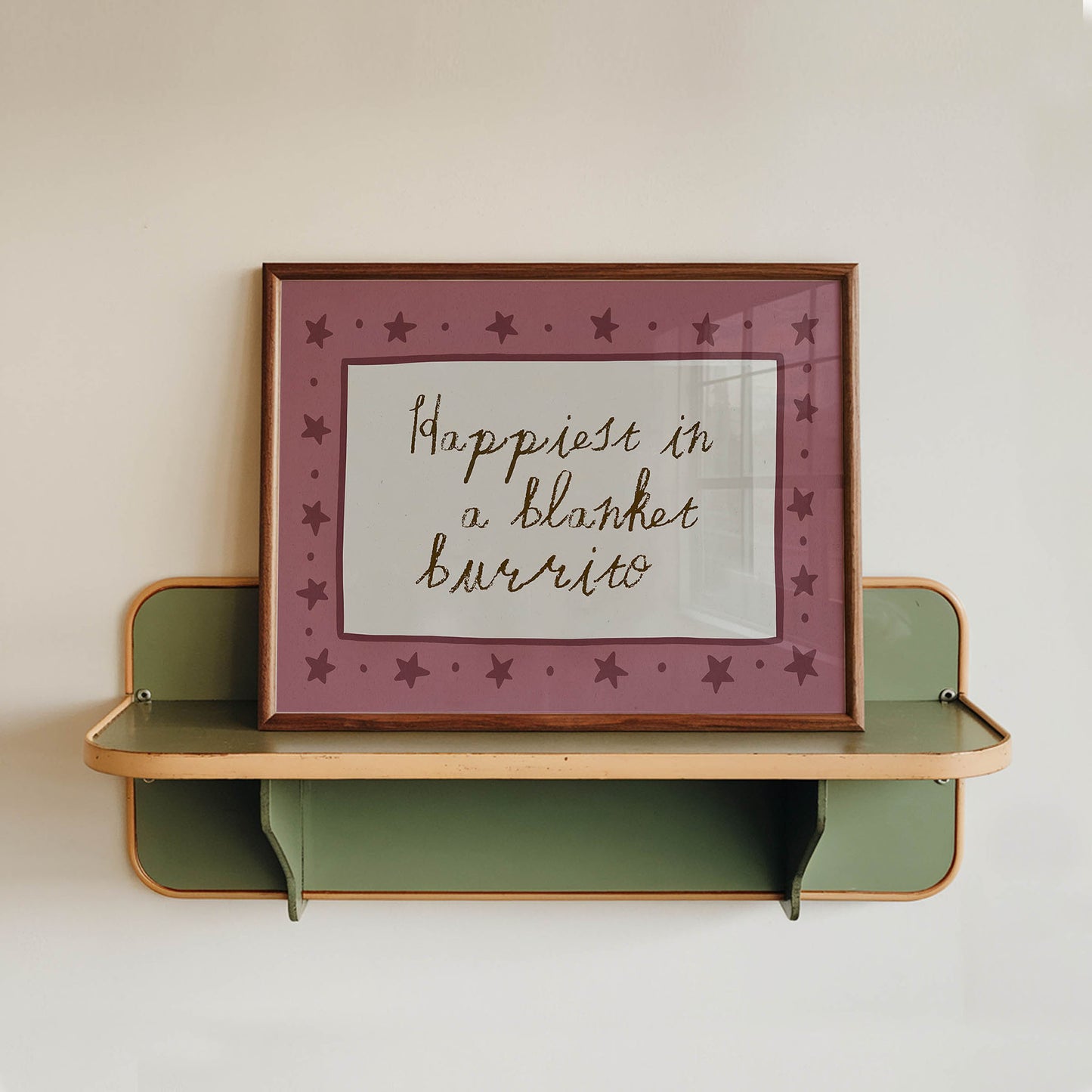 A framed picture with a pink border and purple stars is hanging on a white wall above a green shelf.