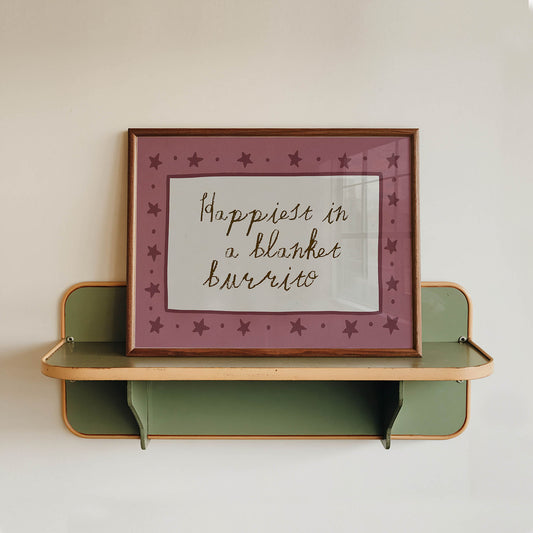A framed picture with a pink border and purple stars is hanging on a white wall above a green shelf.
