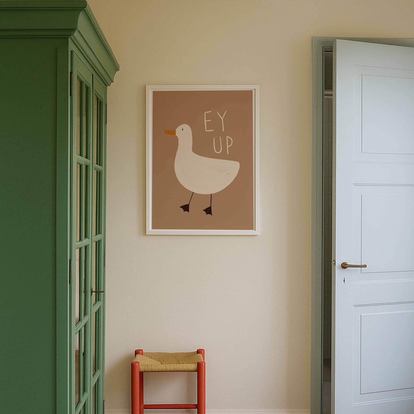 A framed illustrated, fun wall art print of a duck with the yorkshire phrase 'ey yp' written above it on a light brown background. Hanging in a white frame on a wall by a green cupboard and a door with a red chair below it.
