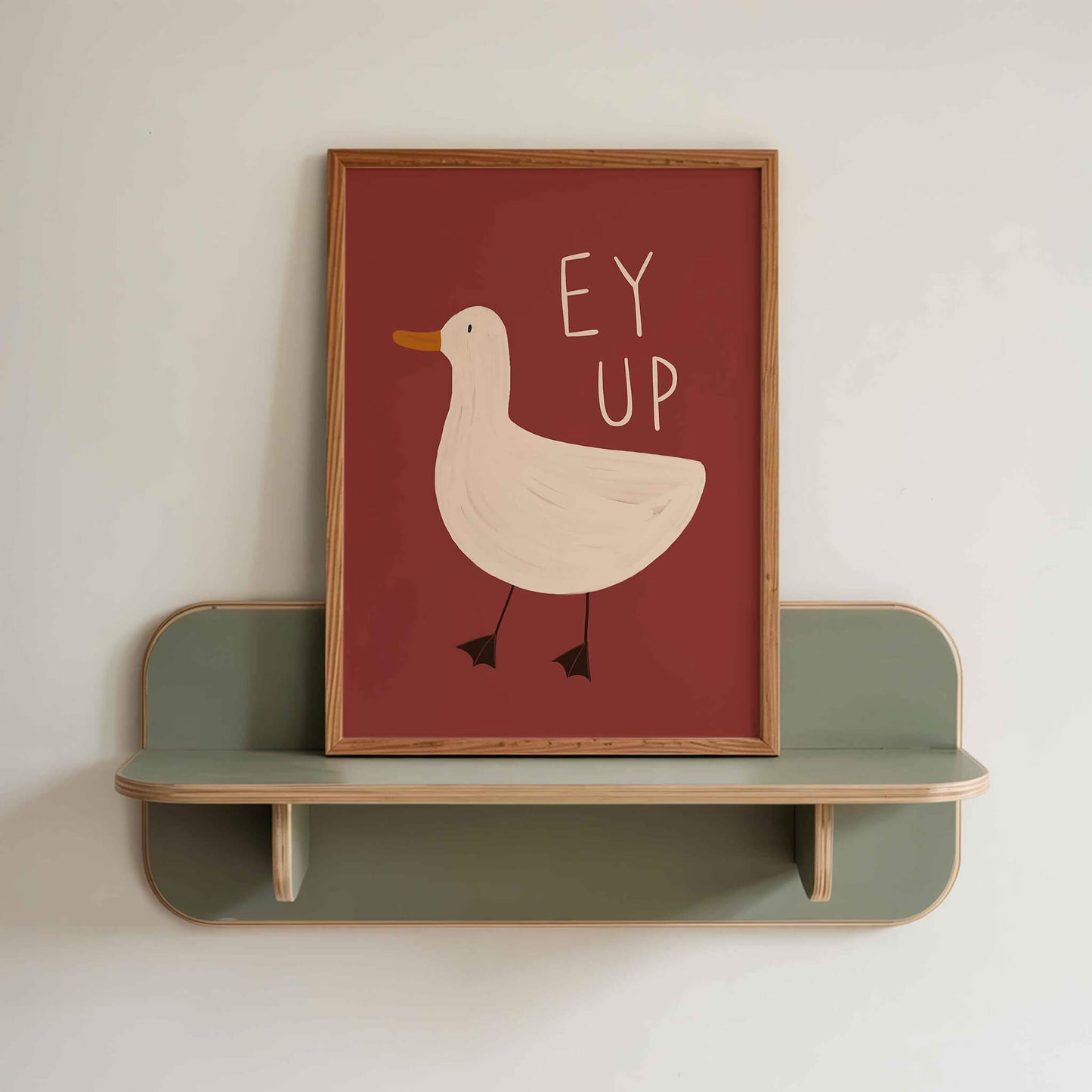 Quirky framed wall art with a burgundy background and a duck illustration with the yorkshire phrase 'Ey Up'. Sitting on a green retro shelf against a white wall.