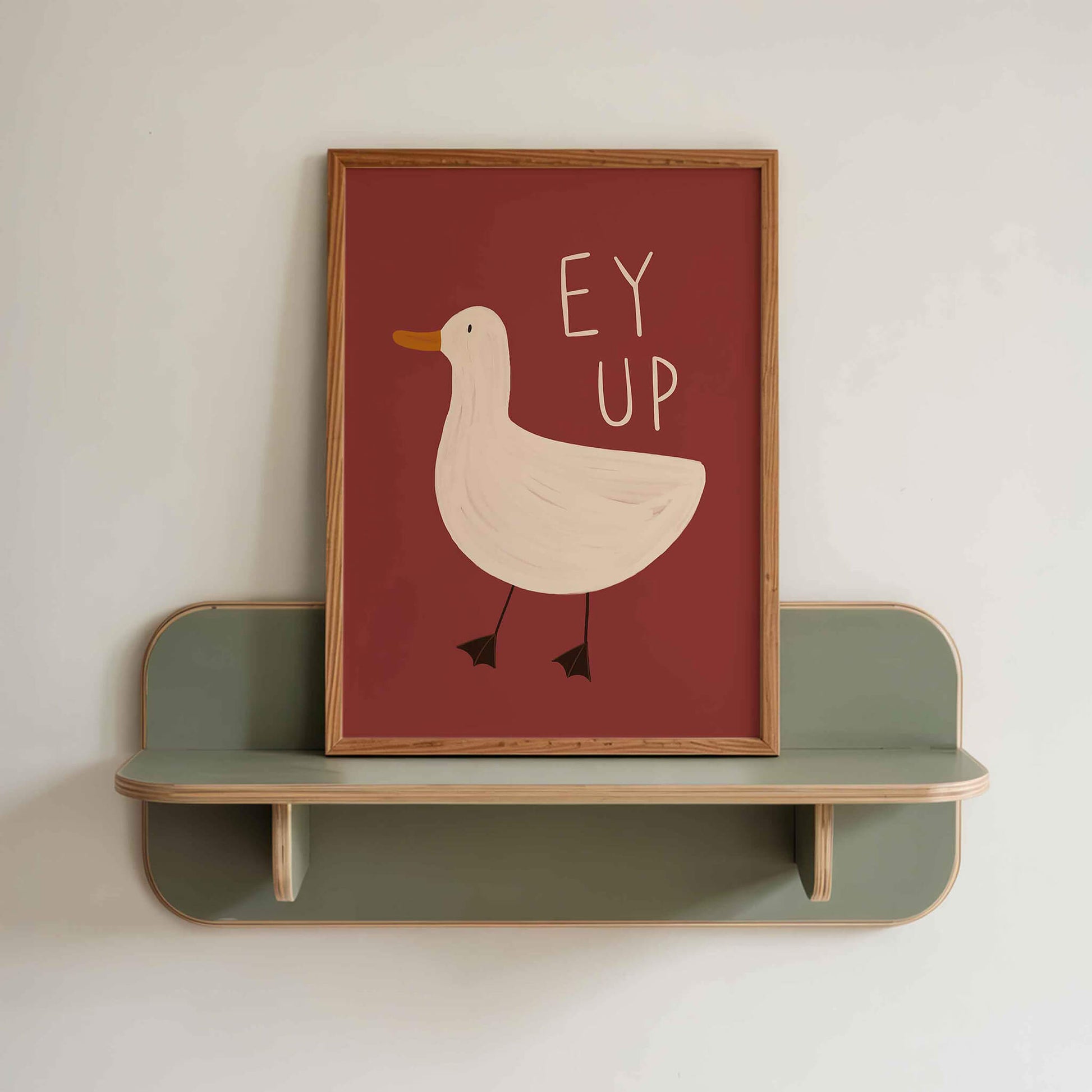 Quirky framed wall art with a burgundy background and a duck illustration with the yorkshire phrase 'Ey Up'. Sitting on a green retro shelf against a white wall.