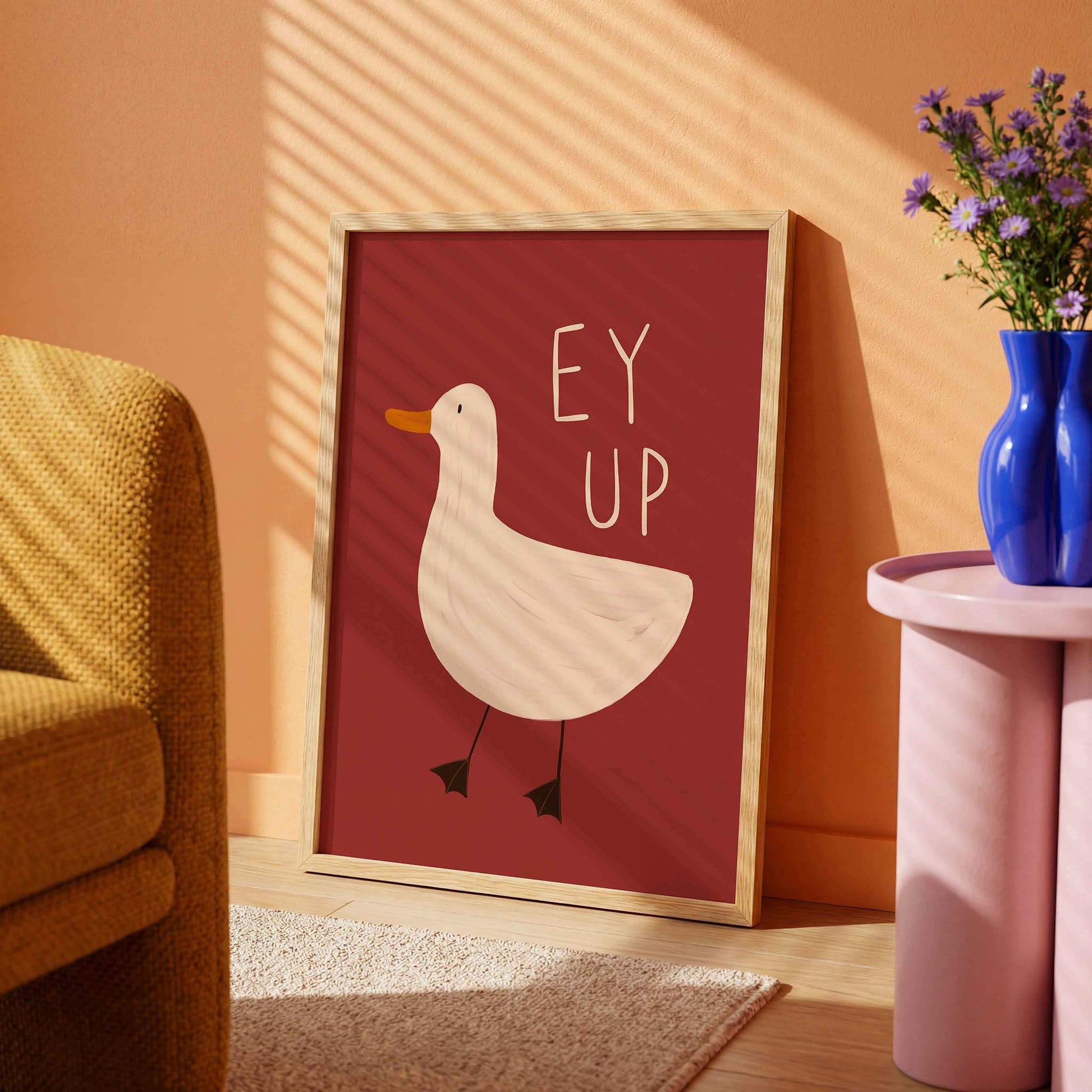 A fun, illustrated, colourful framed print in a dopamine decor themed room with an orange wall, yellow chair, pink table and blue vase with purple flowers in. The print has a burgundy background and a hand drawn picture of a duck with the Yorkshire slang 'ey up' written above it.