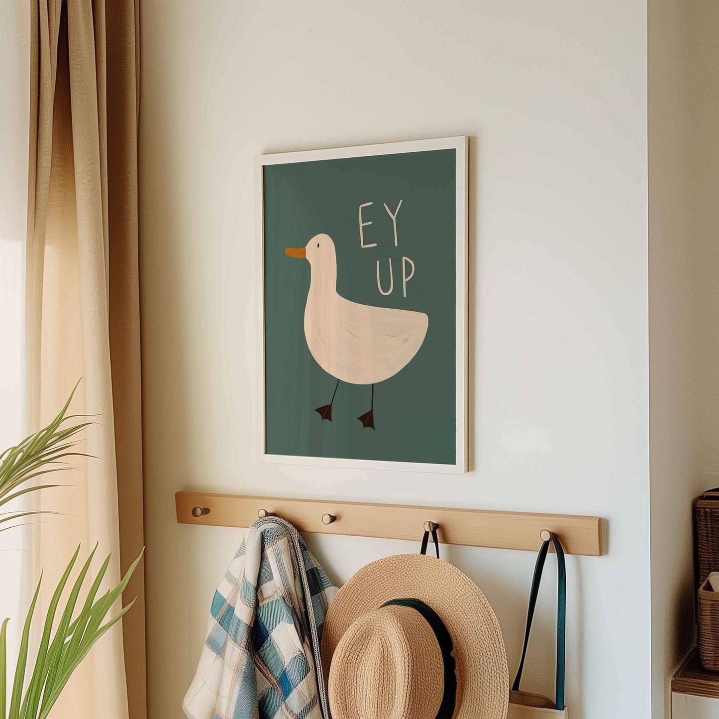 Framed artwork with a duck illustration and 'Ey Up' written on a teal green background on a wall above a wooden hook with hats and a coat.