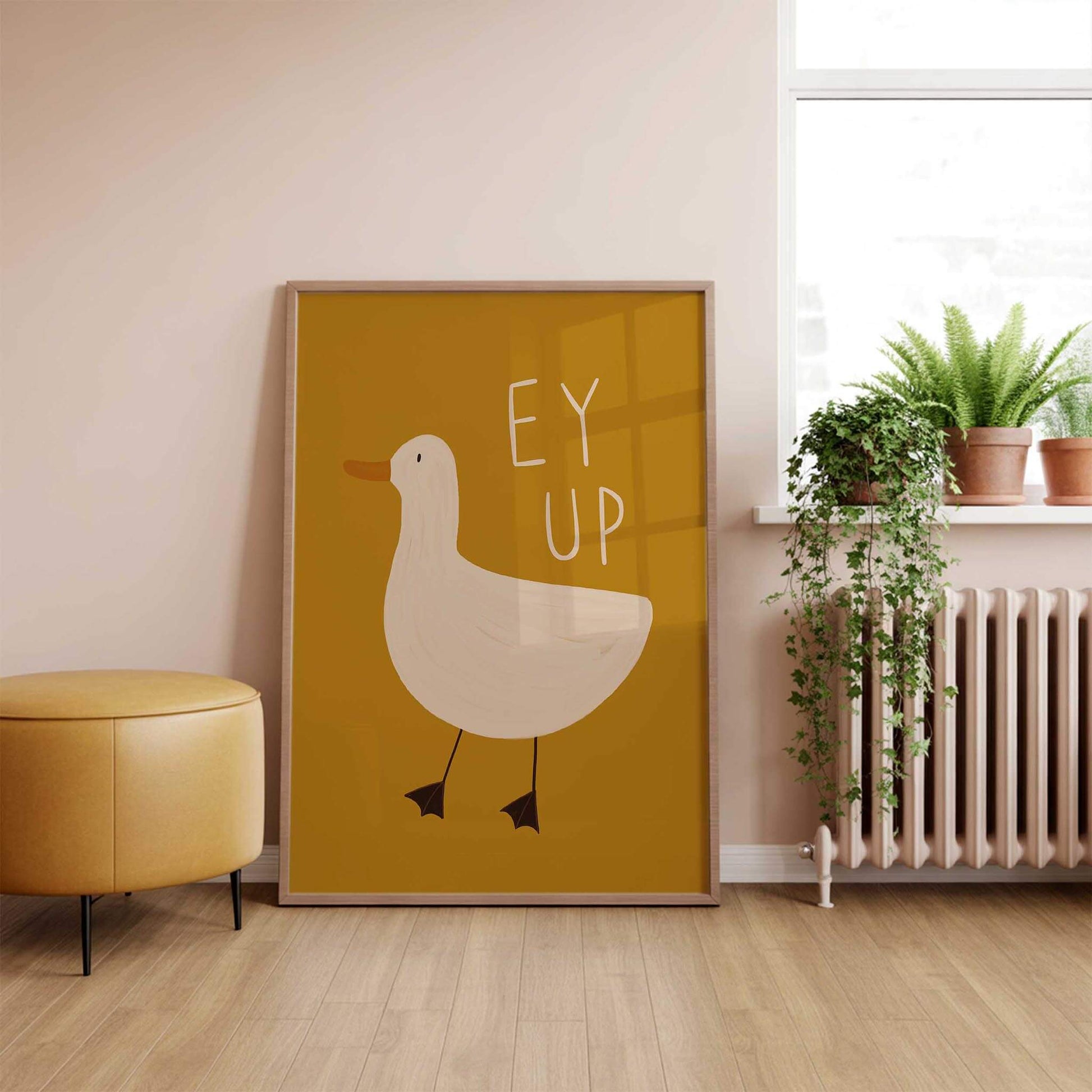 A Dandy Prints framed large artwork of a hand illustrated duck on a mustard yellow background with the Yorkshire slang 'EY UP' written above the duck. In a living room by a windowsill with plants and a yellow footstool.